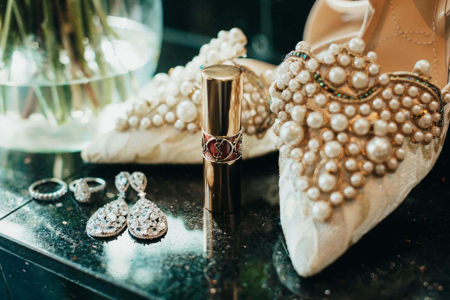 Wedding accessories including pearl-adorned shoes, jewelry and lipstick arranged on reflective surface.