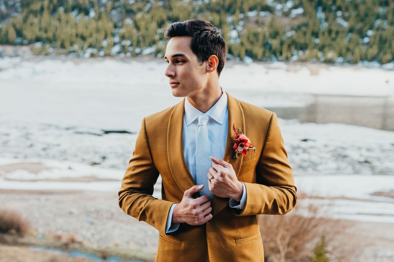 Groom in camel suit and light blue tie poses against winter mountain backdrop.