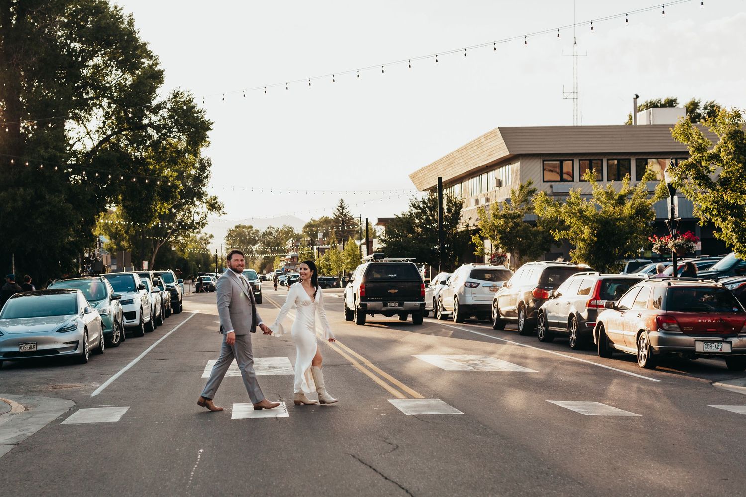 Wedding couple crossing street in formal attire with parked cars in background.