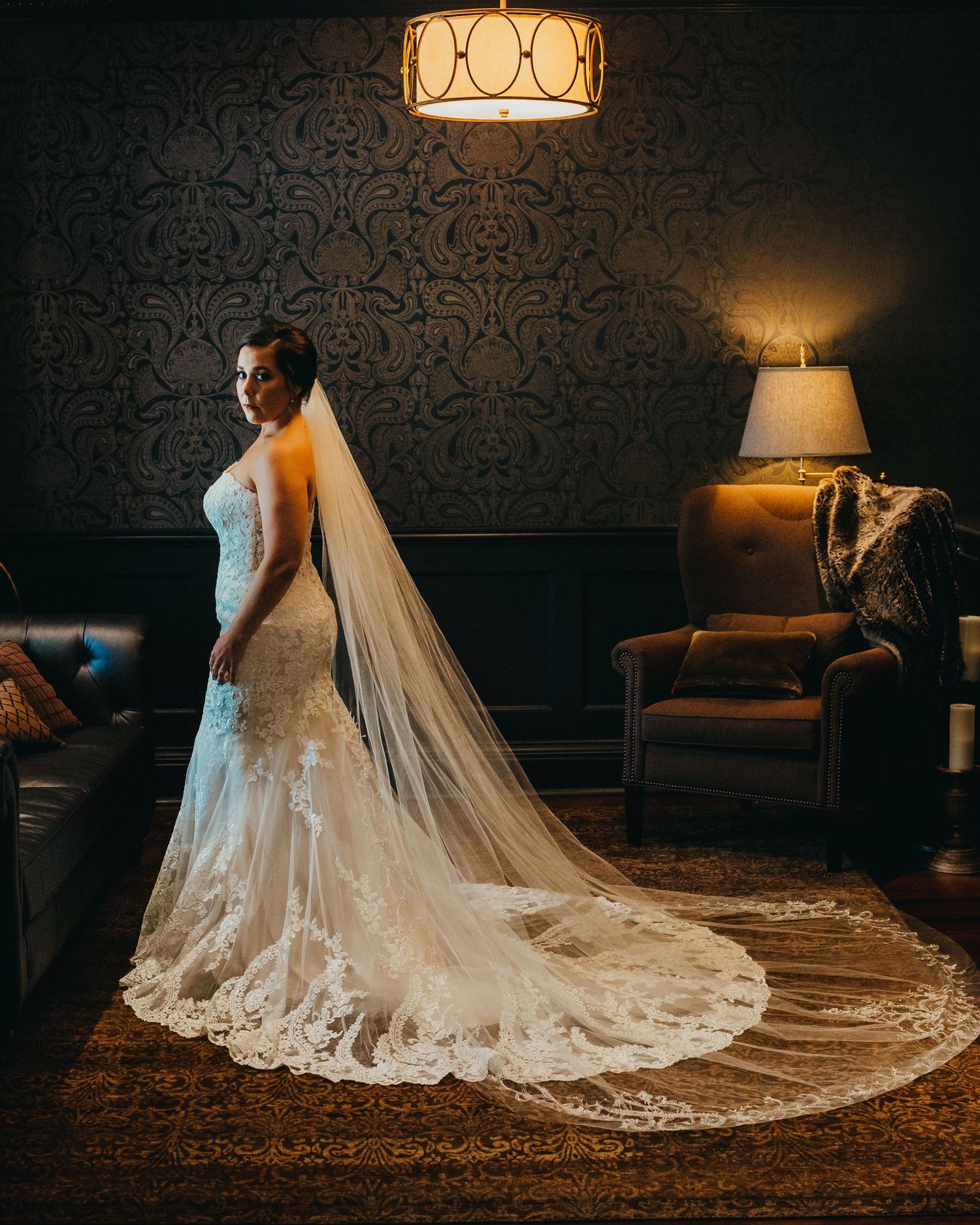 Elegant bride in lace wedding dress with cathedral veil posing against dark damask wallpaper.