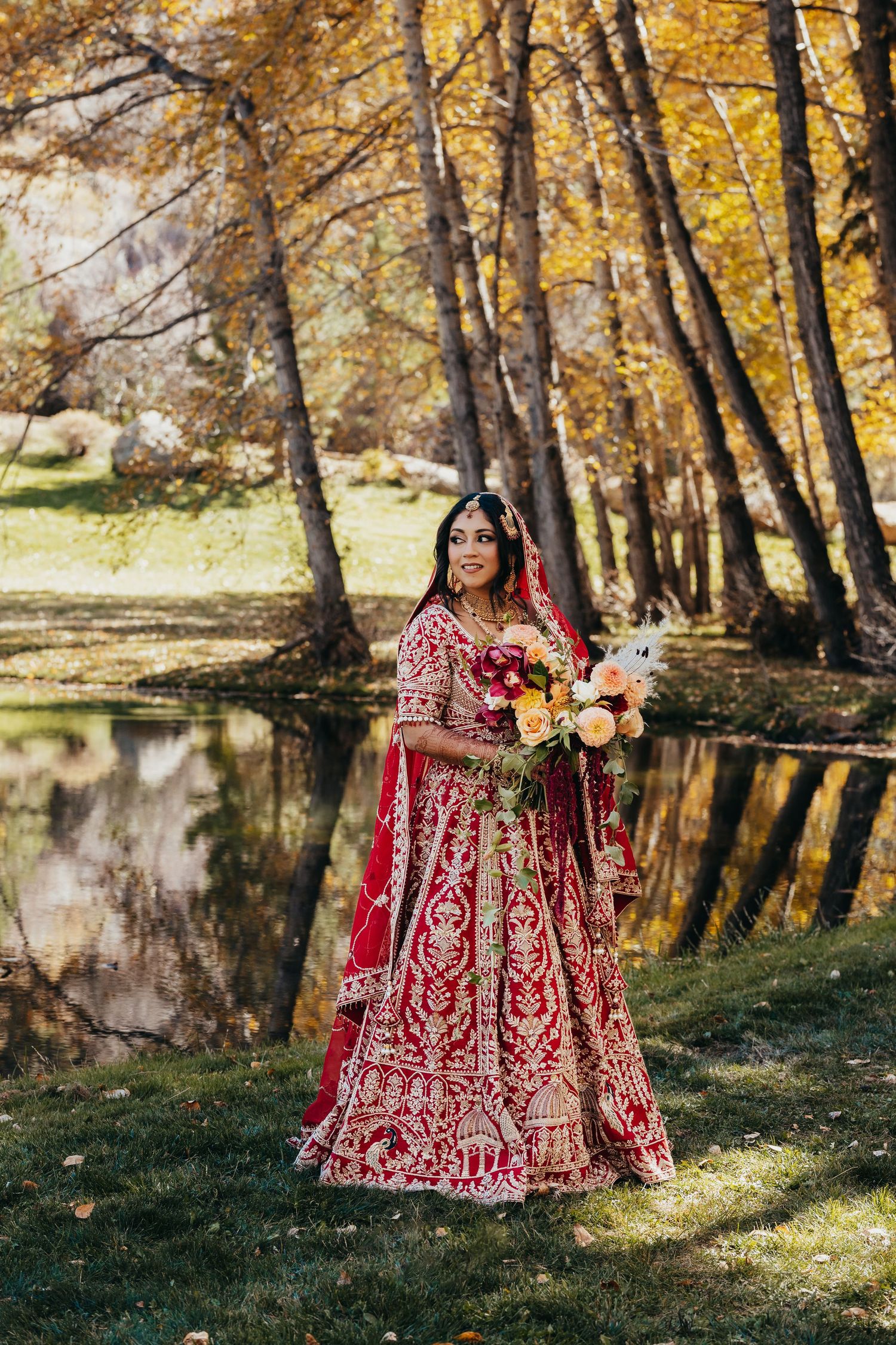 Bride in ornate red and gold lehenga holds bouquet in autumn forest setting.