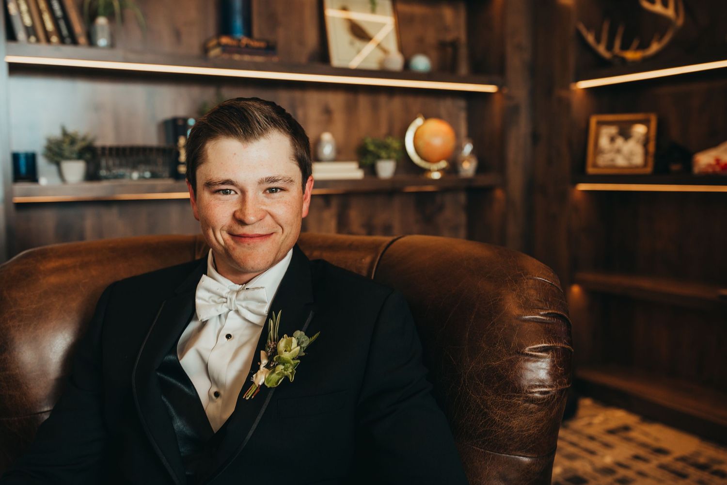 A groom in a dark suit and tie sits on a brown leather chair smiling in a rustic wood-paneled room.
