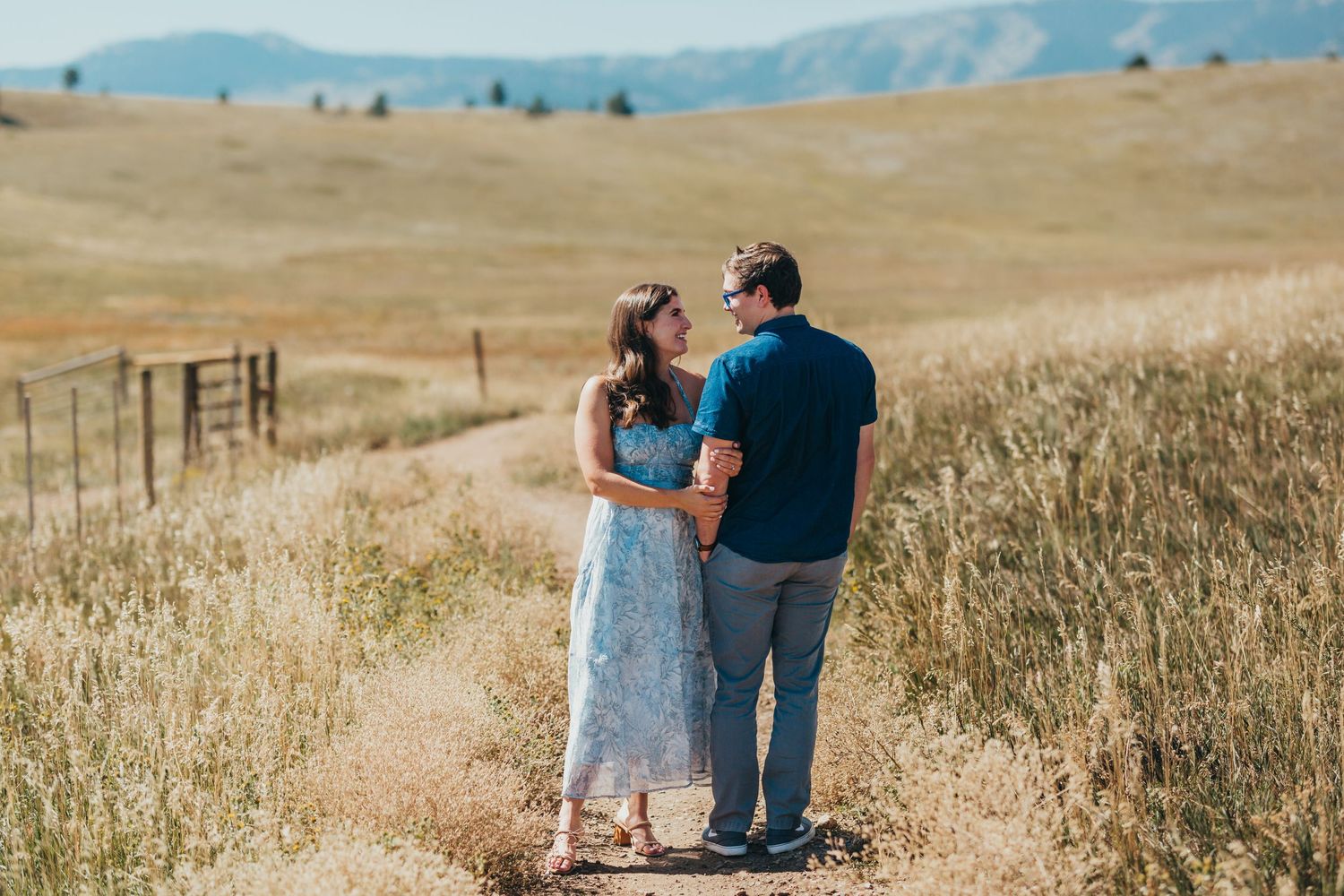 Couple standing on dirt path overlooking rolling hills and mountains in distance.