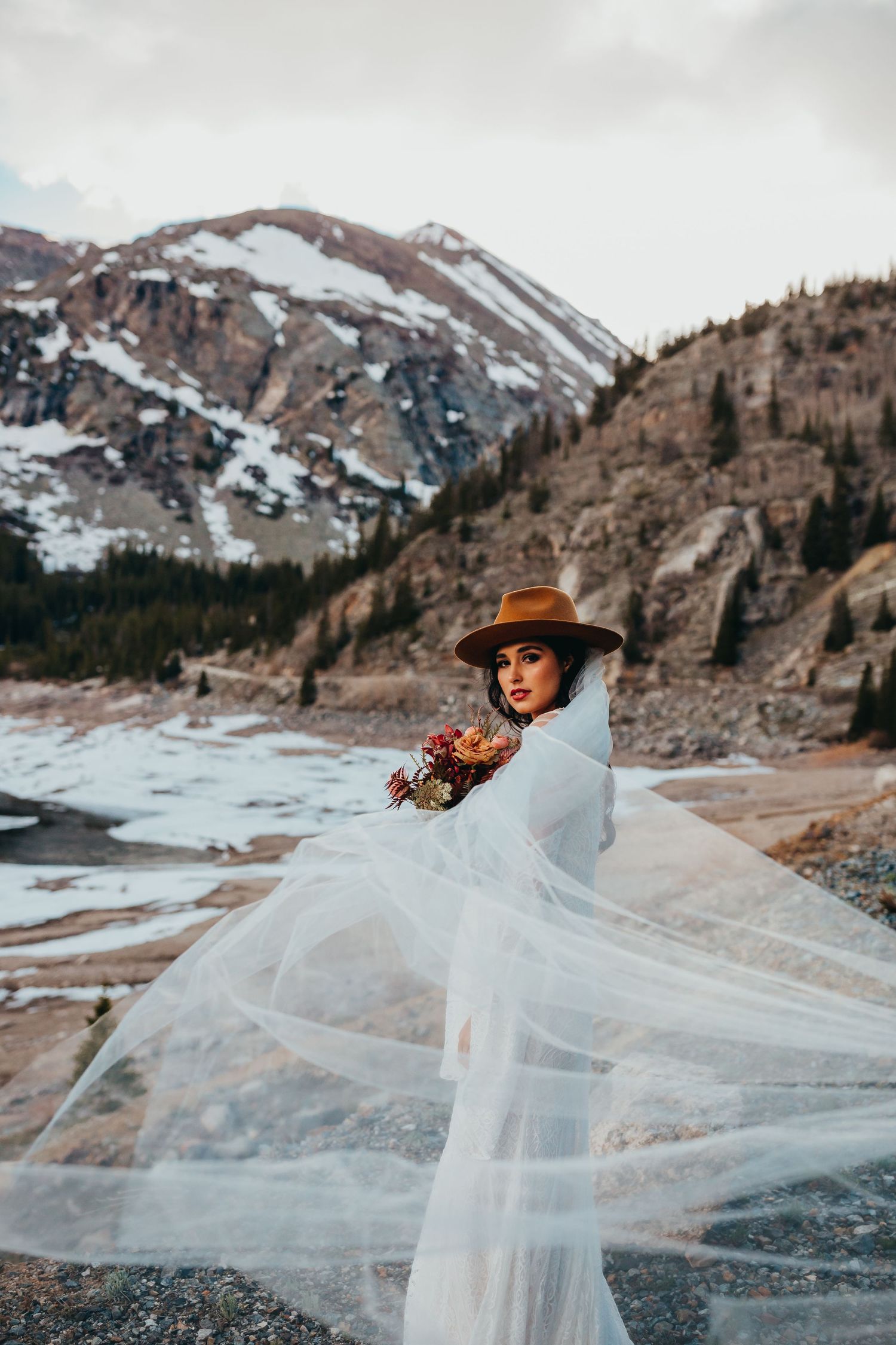 Wedding portrait with billowing veil against snow-capped mountains and frozen lake.