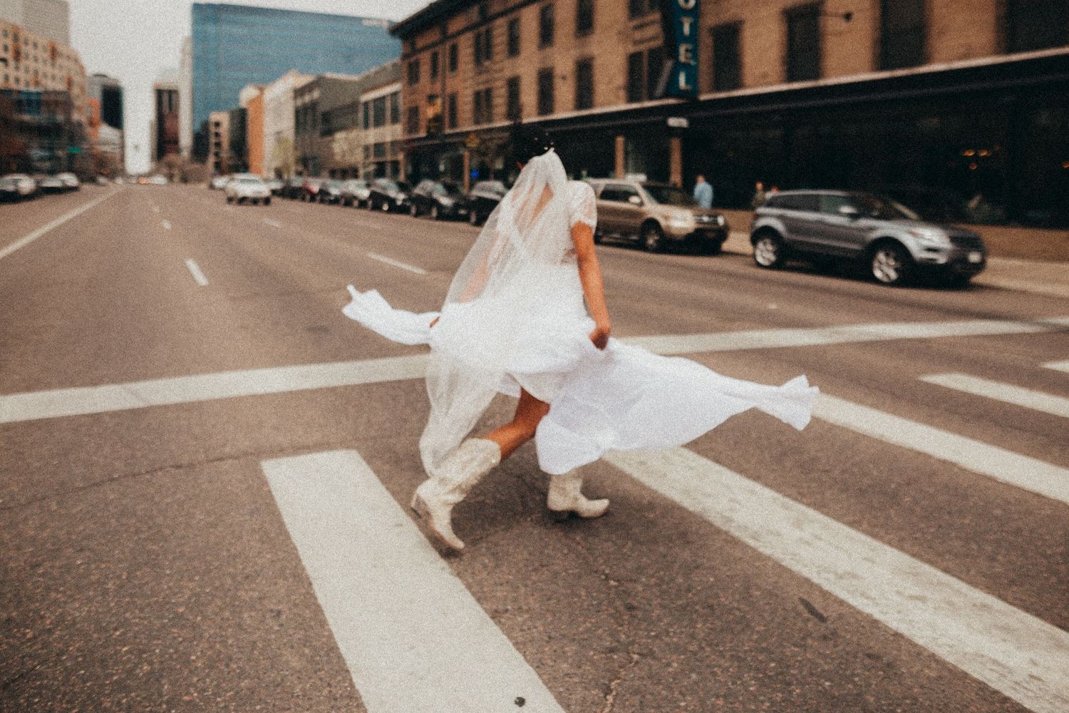 A bride in a white dress crosses an urban street intersection with flowing fabric.