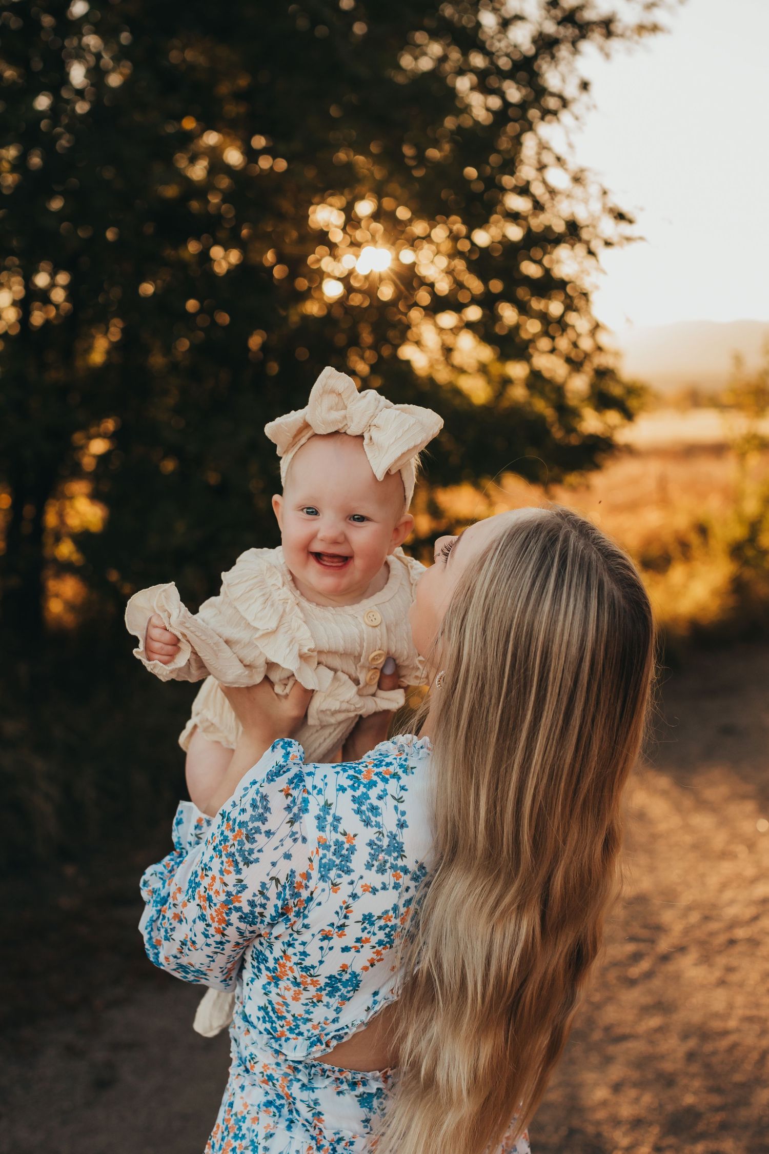 Sunset portrait of mother holding baby wearing cream-colored outfit with bow.