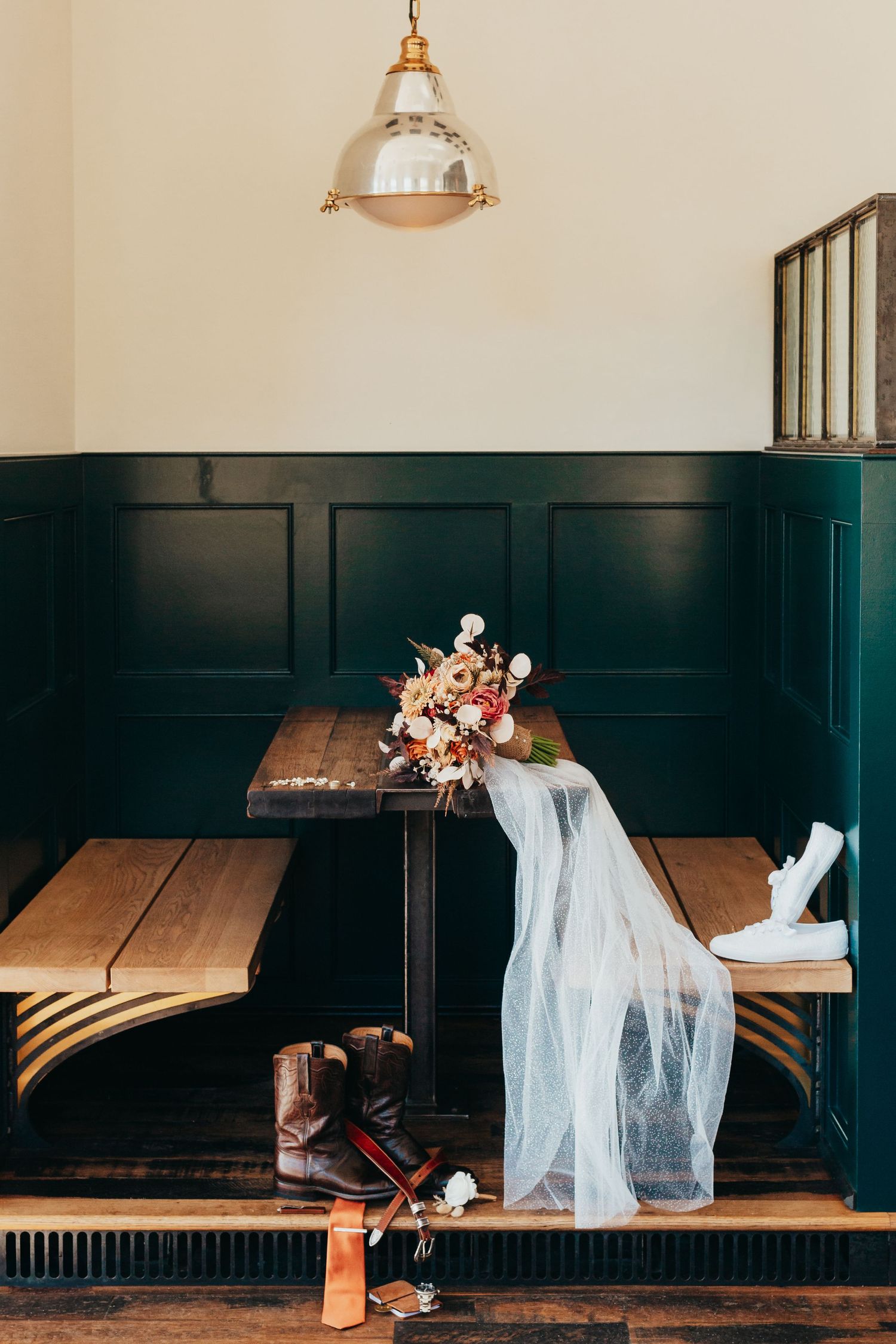 Wedding details with floral arrangement and veil on wooden bench under pendant light.