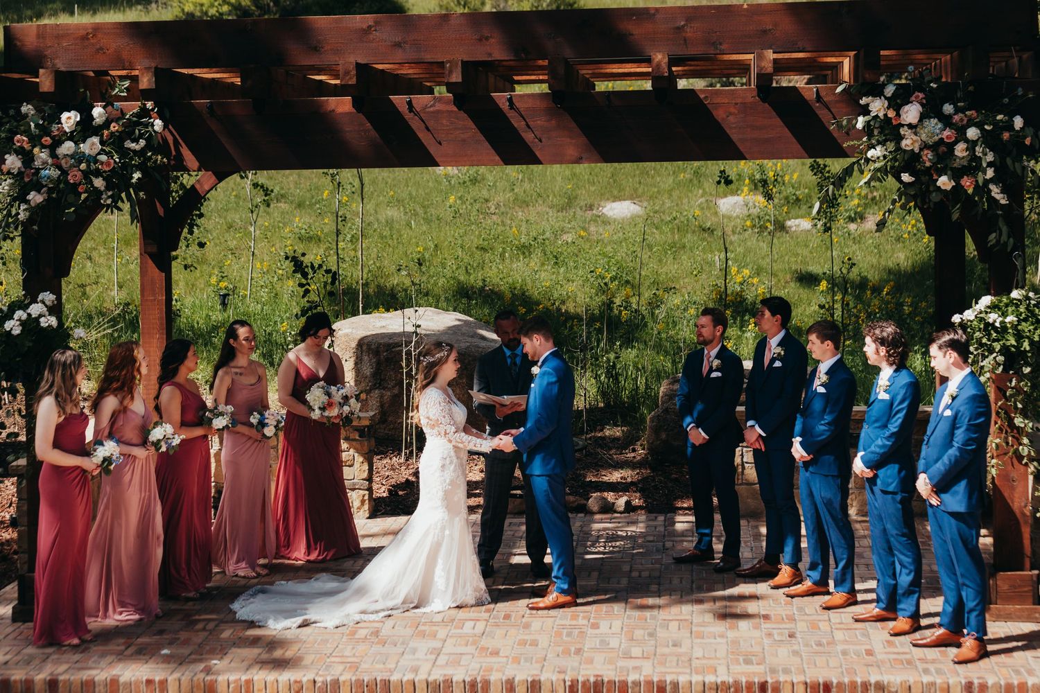 Wedding ceremony under wooden pergola with pink-dressed bridesmaids and groomsmen in blue suits.