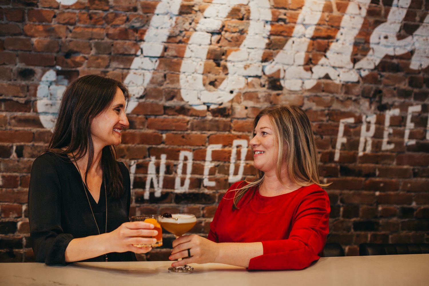 Two friends share drinks and conversation at a casual cafe with rustic brick wall backdrop.