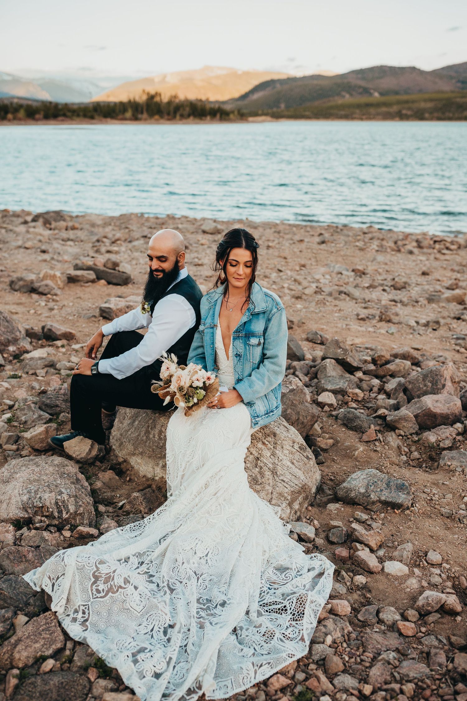 Wedding couple in white dress and denim jacket sitting on rocky lakeshore.