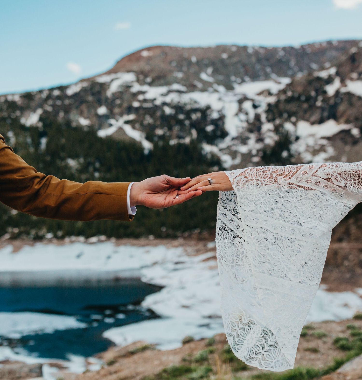 A hand holds a delicate lace veil against a snowy mountain backdrop with a blue alpine lake.