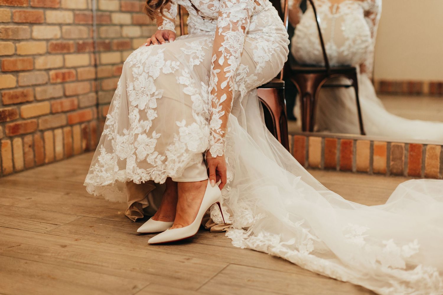 Detail shot of white lace wedding dress hem and bridal shoes against brick wall and wooden floor.