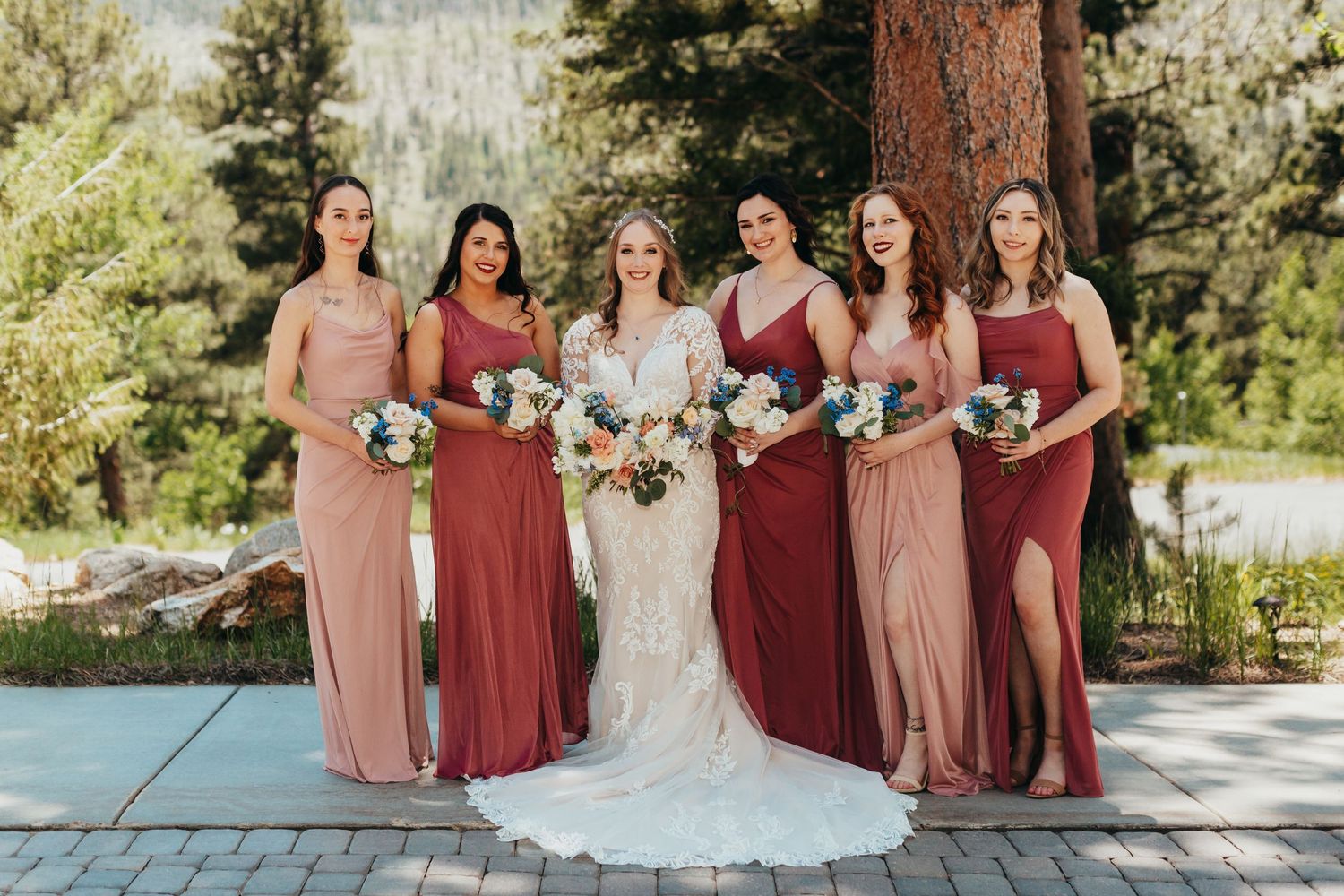 Bridesmaids in varying shades of red and pink dresses pose with bouquets outdoors.