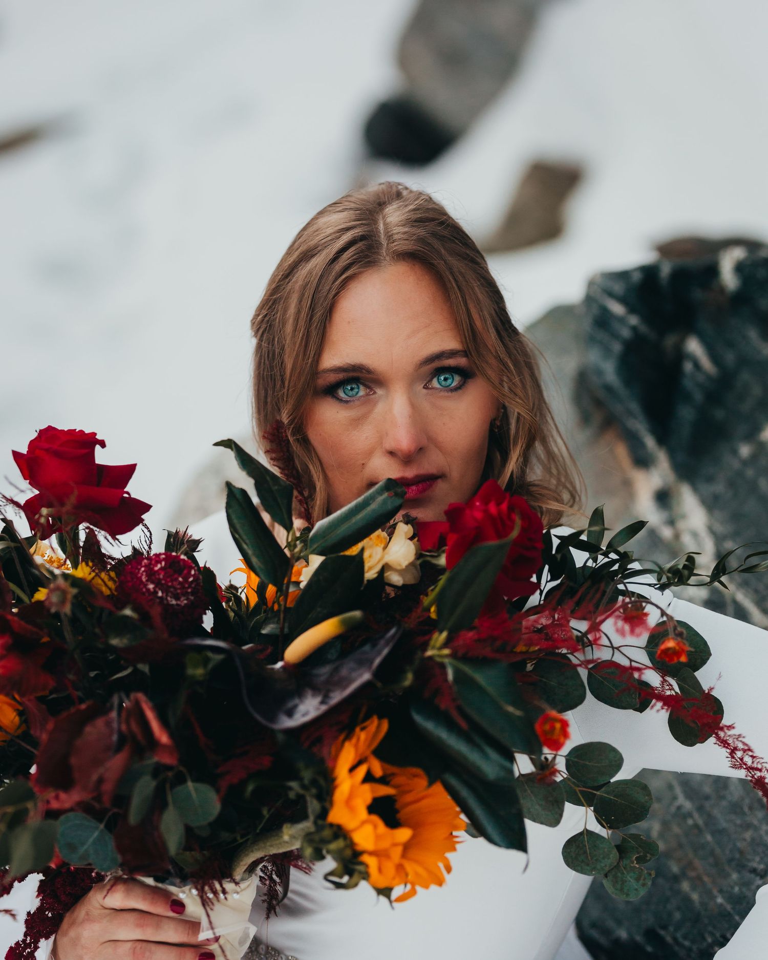 A vibrant winter bridal bouquet with deep red roses and sunflowers held against a snowy backdrop.