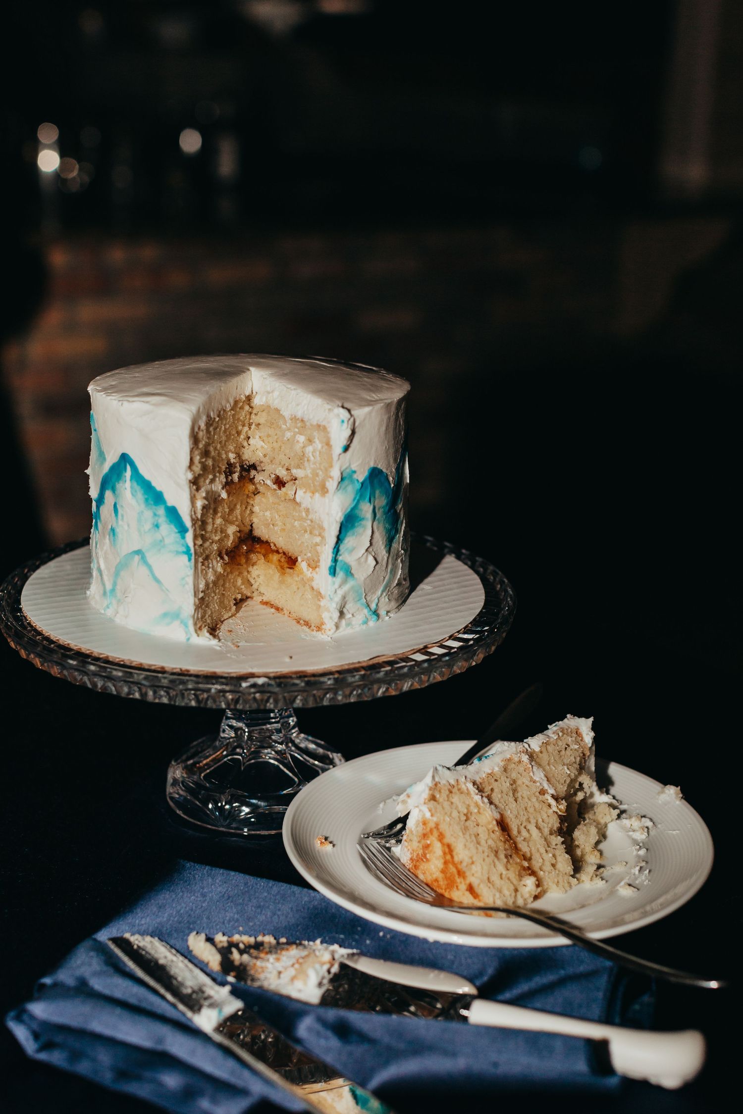 A sliced white wedding cake with blue mountain designs on display with a piece plated beside it.
