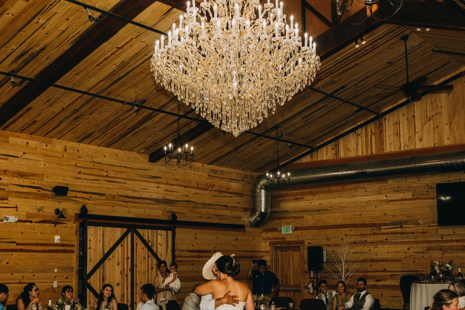 A grand crystal chandelier hangs from wooden beams during a wedding reception celebration.