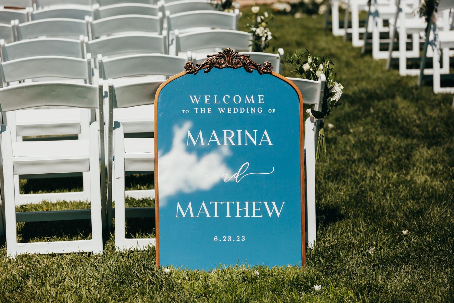 A blue wedding welcome sign sits among white folding chairs on a grassy lawn.