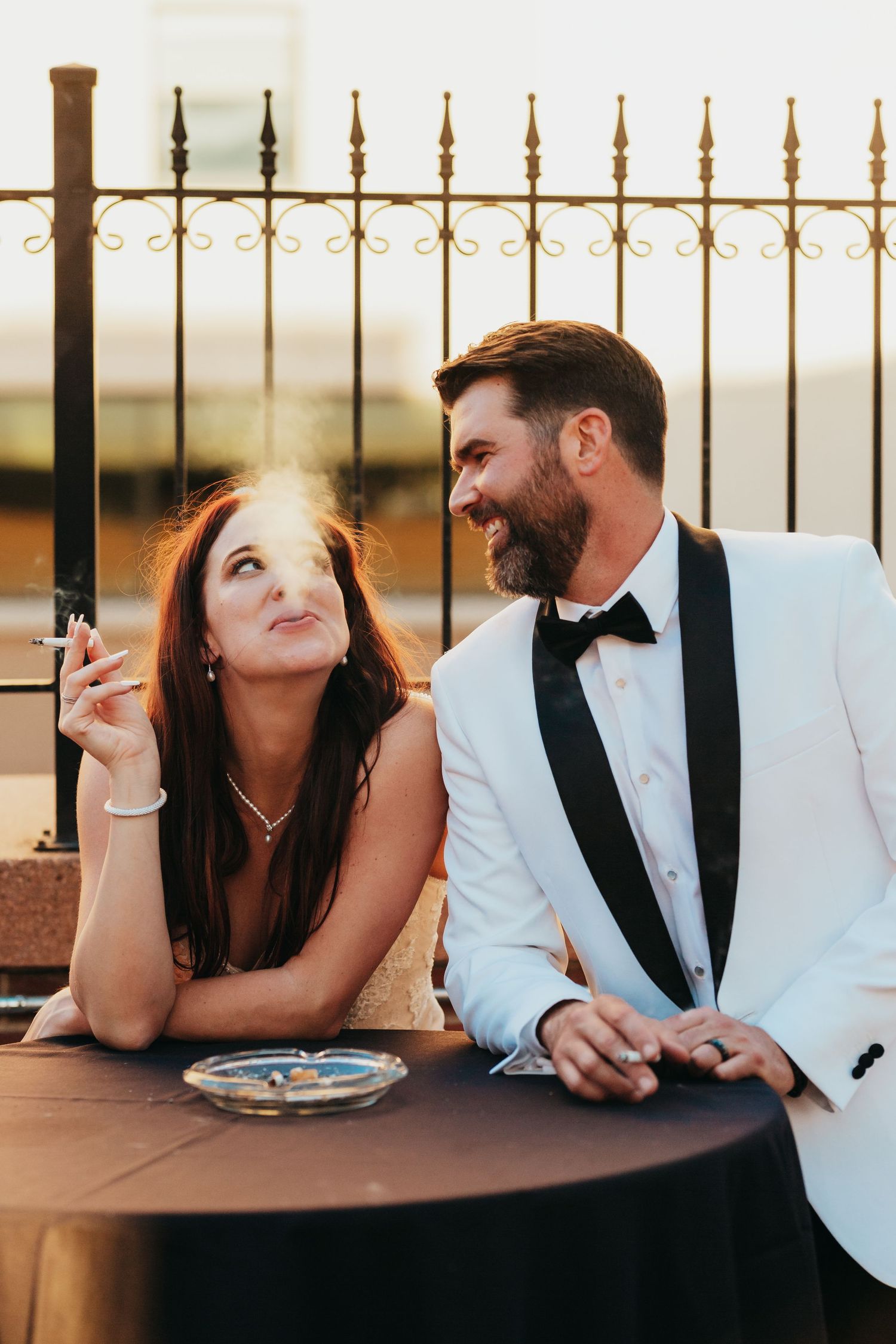 Couple shares intimate moment at outdoor table with decorative iron fence backdrop.