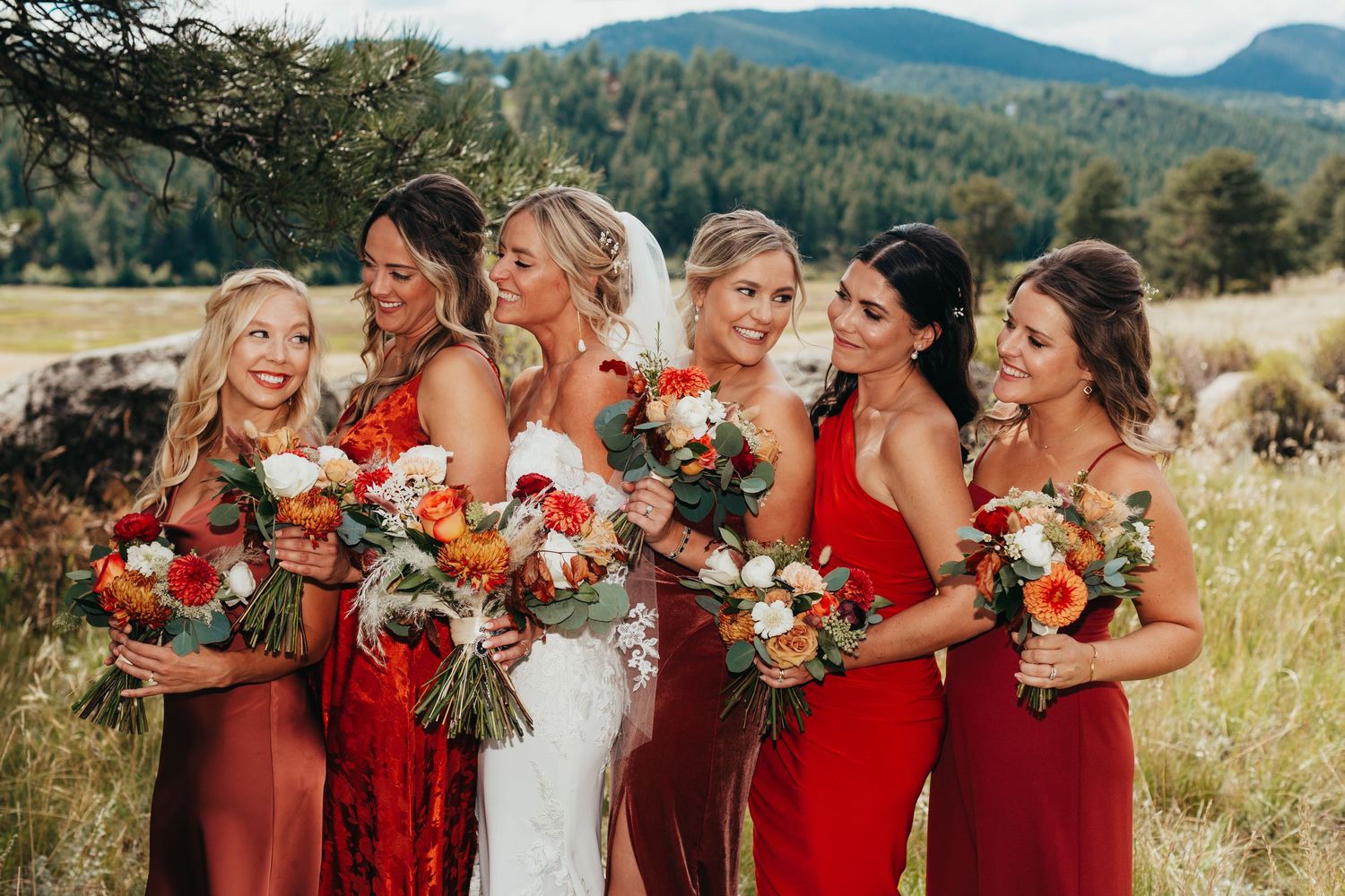 Bridal party in red dresses holding vibrant fall bouquets against mountain landscape.