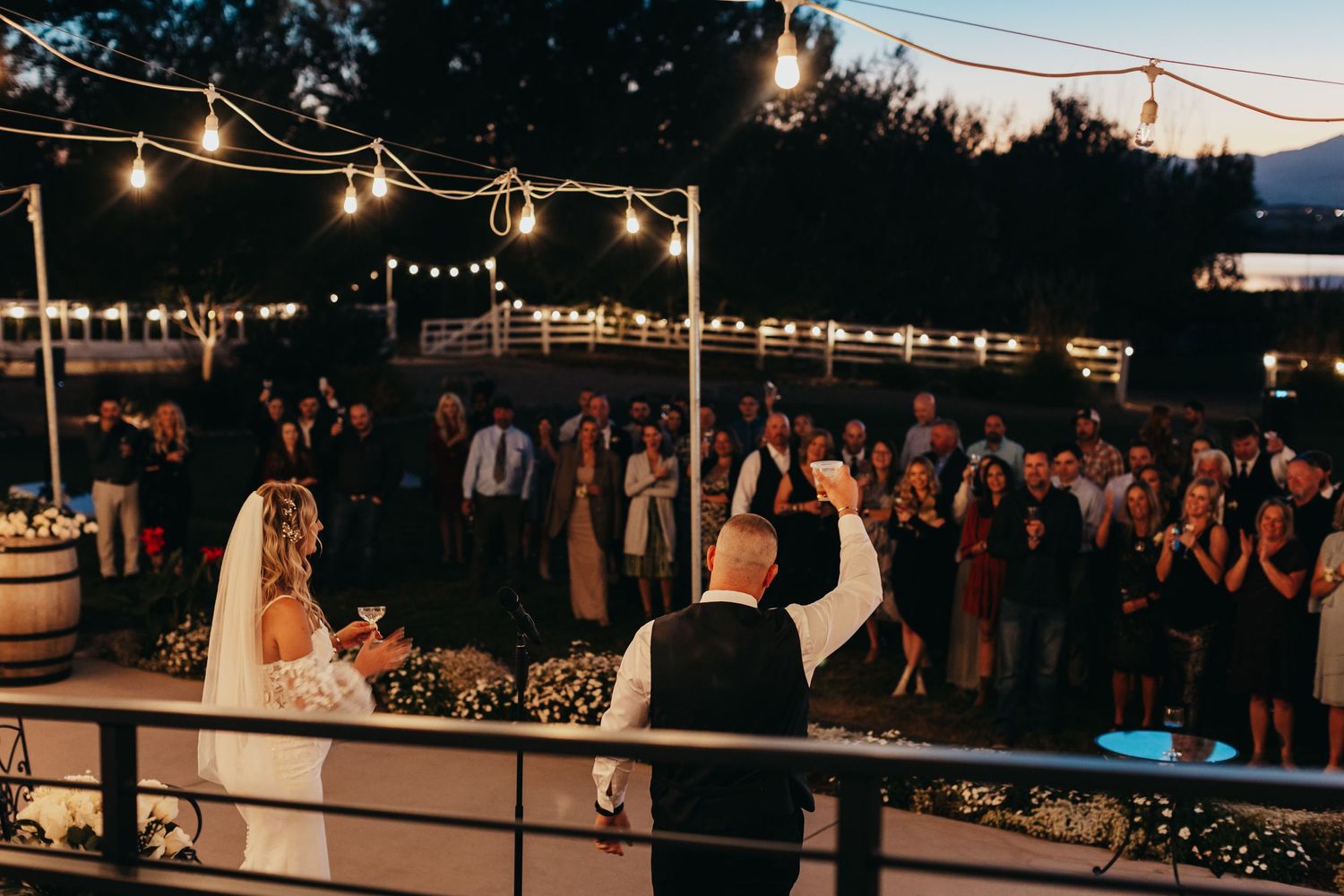 Evening wedding reception with string lights and guests watching couple on outdoor deck.