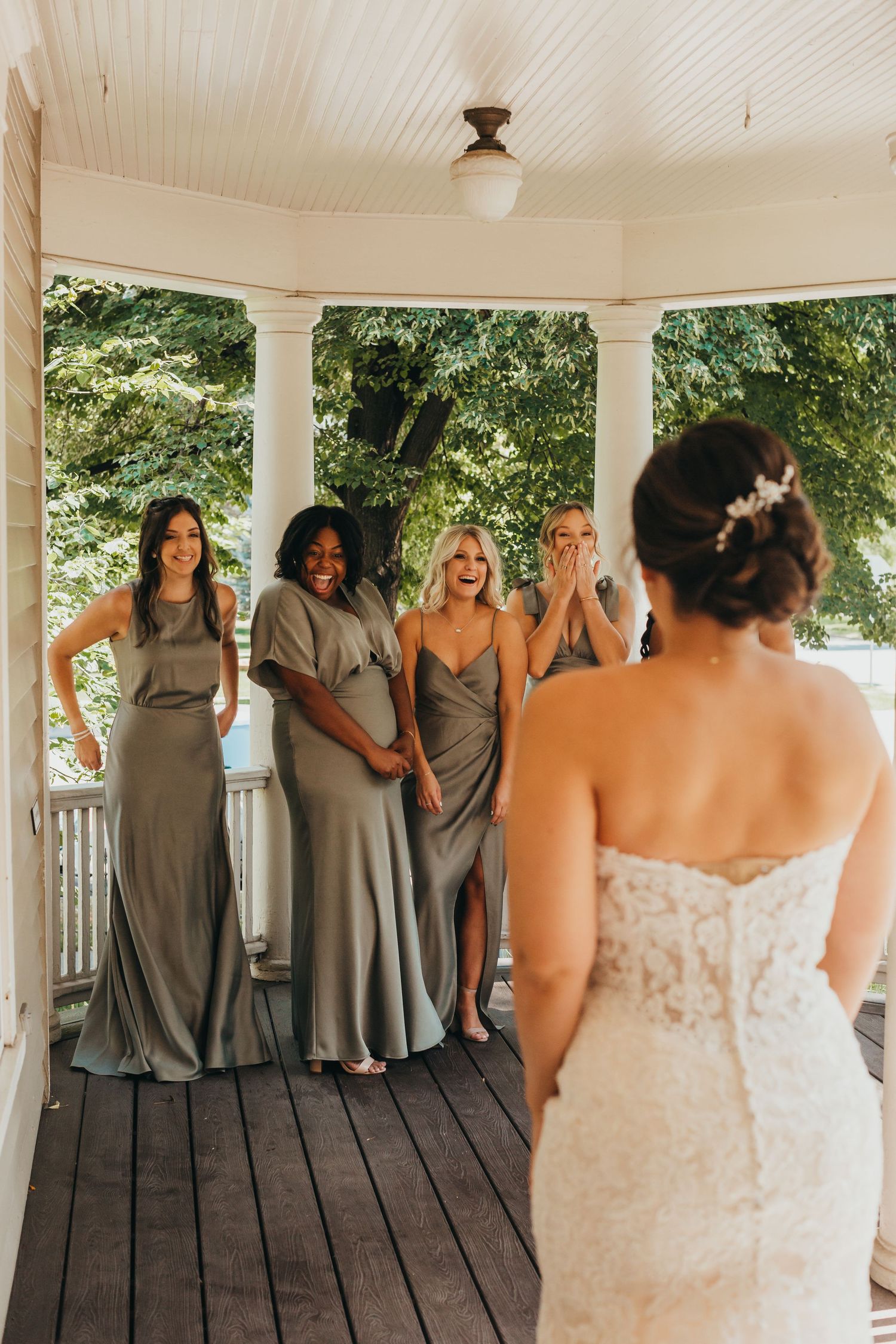 Bridesmaids in flowing grey dresses react emotionally on a covered porch.