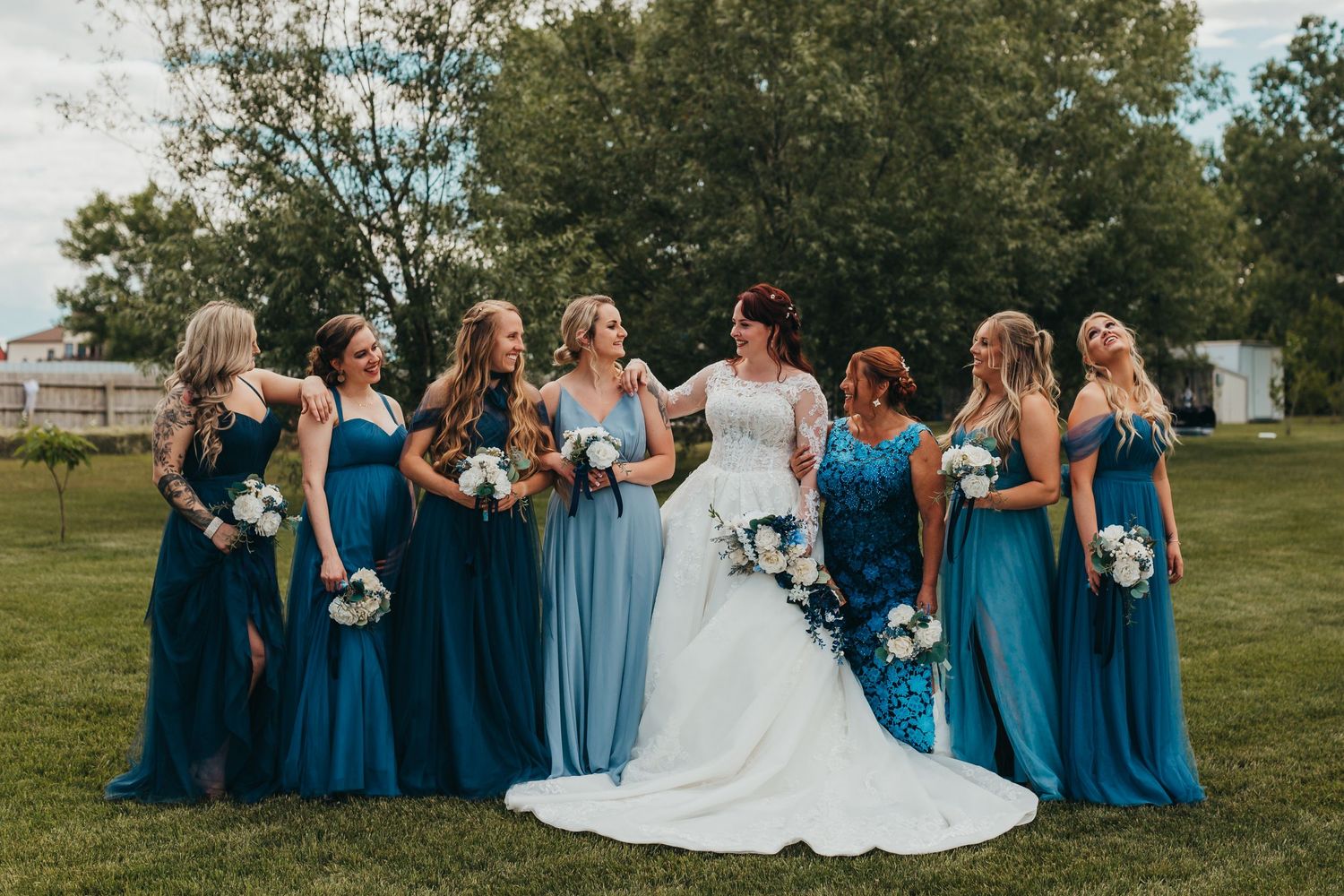 Bridal party in shades of blue dresses pose with bouquets during outdoor wedding.