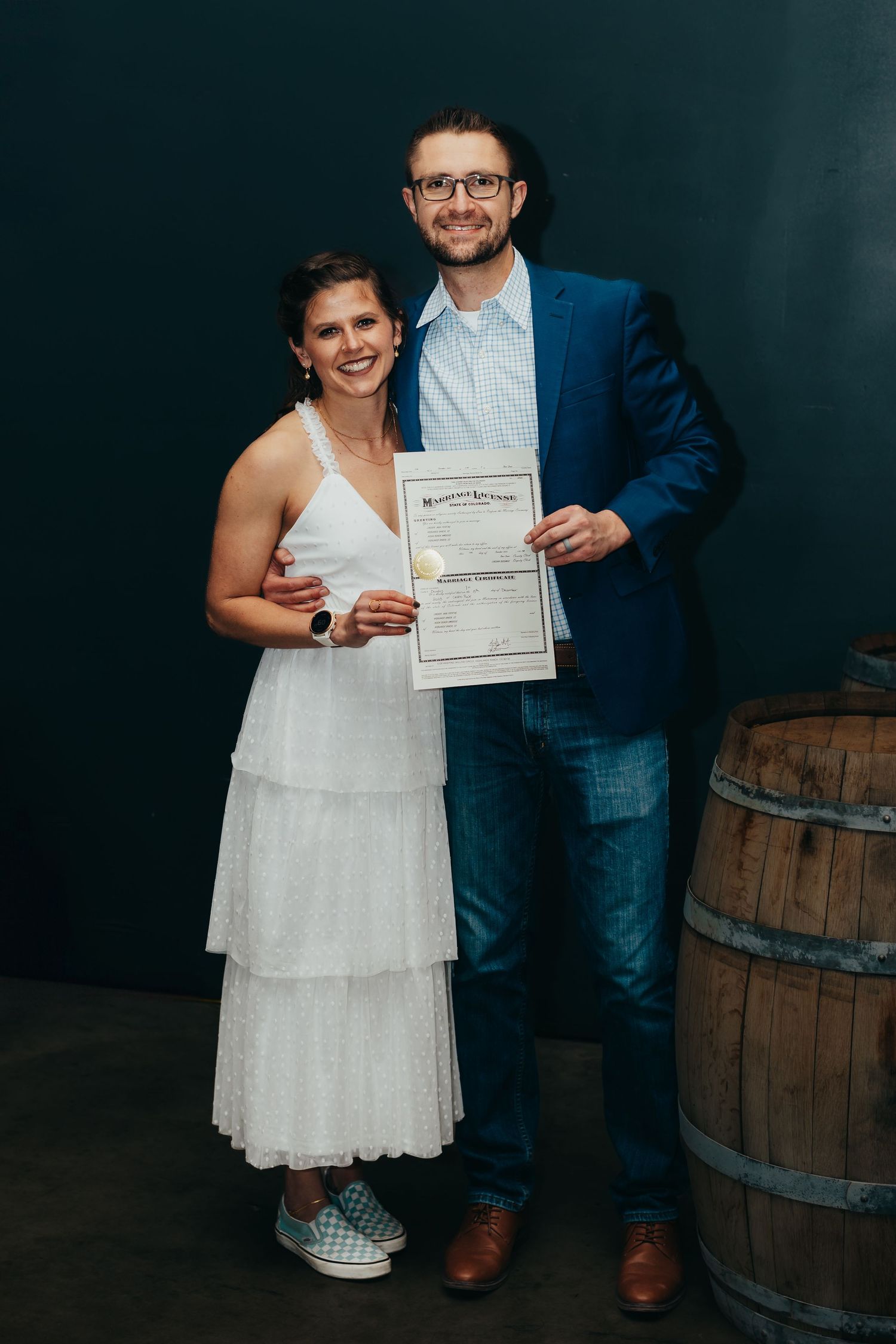 Couple poses with their marriage certificate in front of a wooden barrel and dark backdrop.