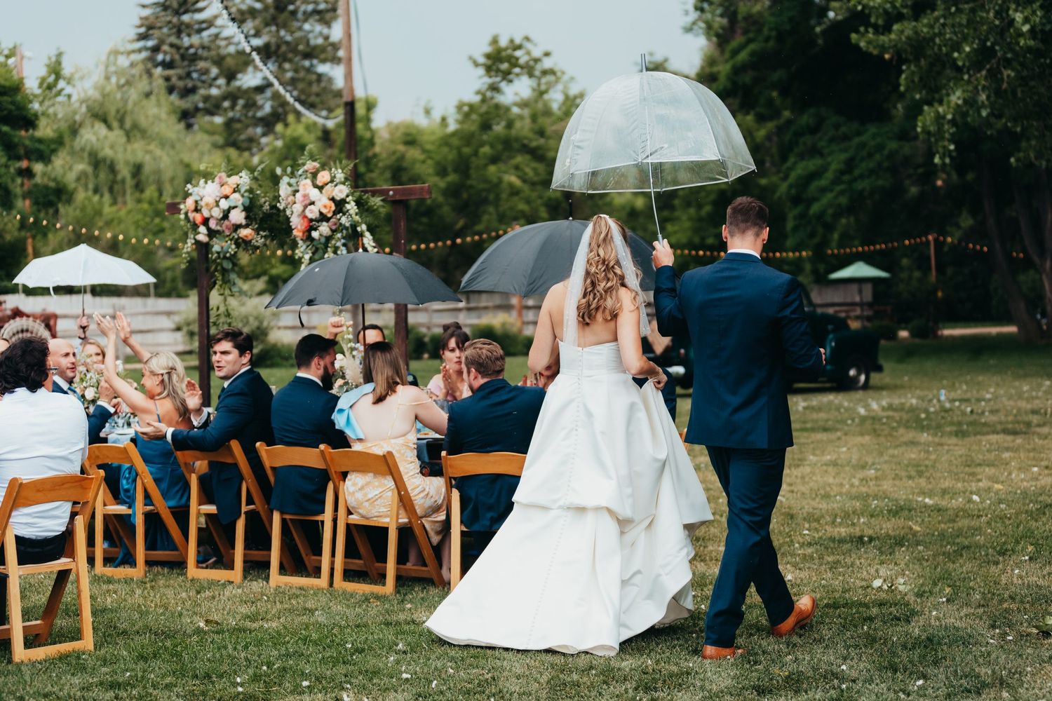 Couple walks down aisle under clear umbrellas during outdoor garden ceremony.