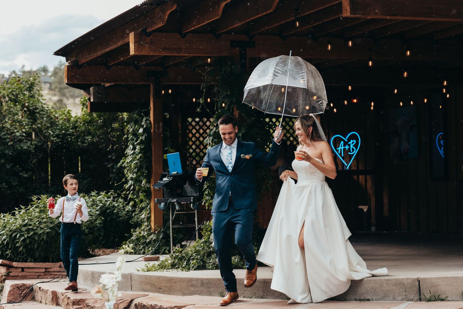 Couple exits ceremony under clear umbrella with string lights and rustic barn in background.