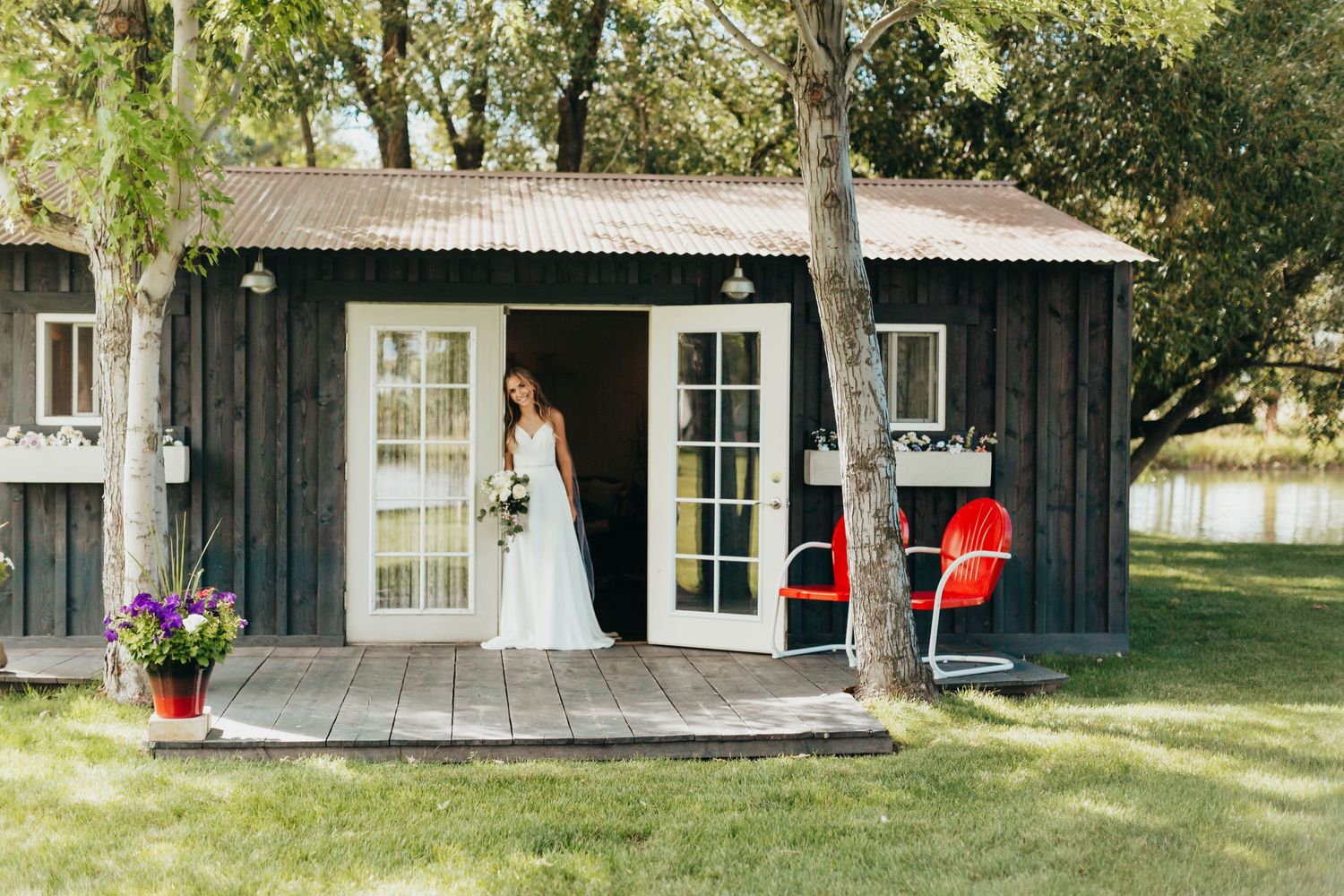 A rustic black cabin with white trim and French doors features a bride standing on the wooden porch.