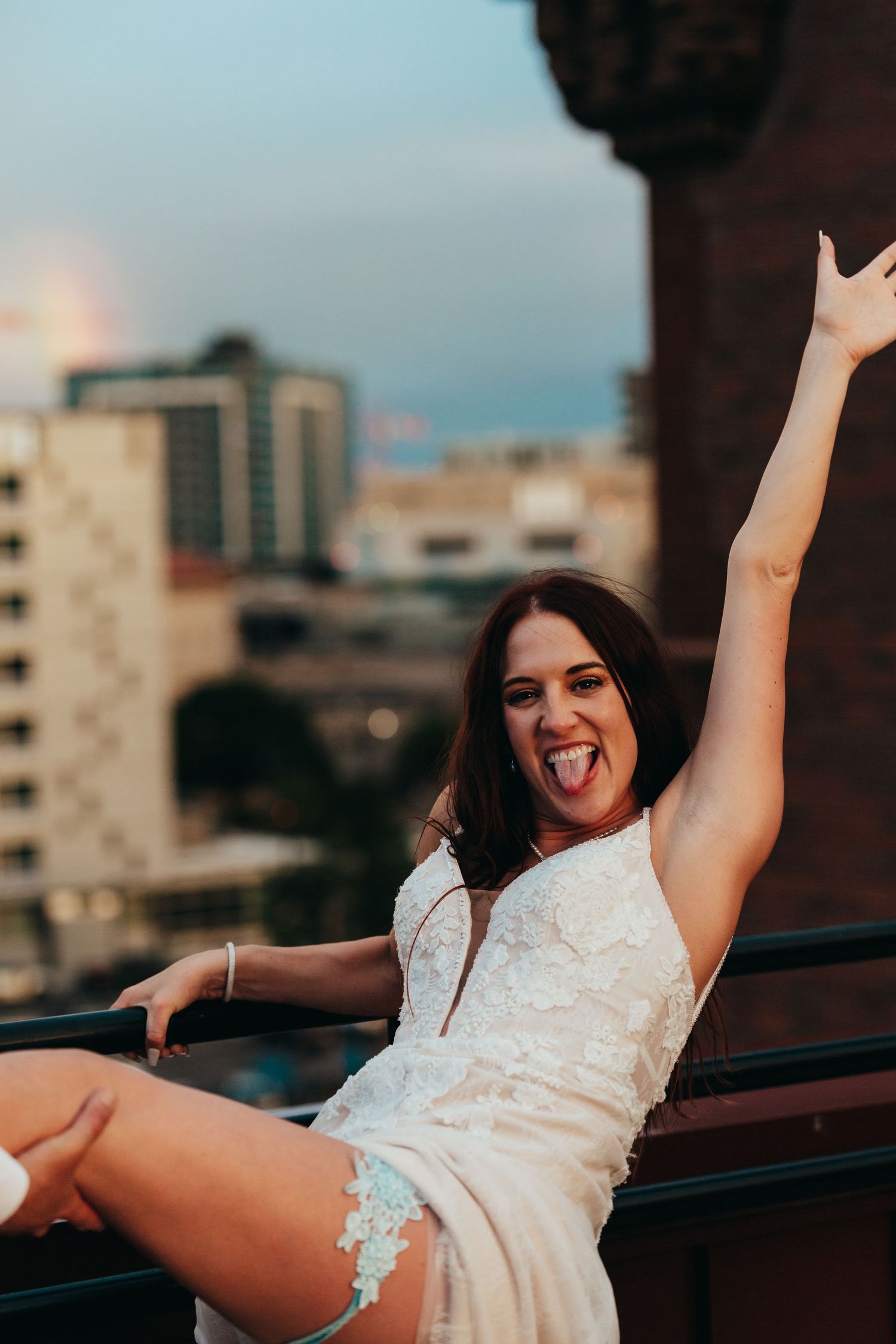 Bride in white dress poses playfully on city rooftop at sunset.