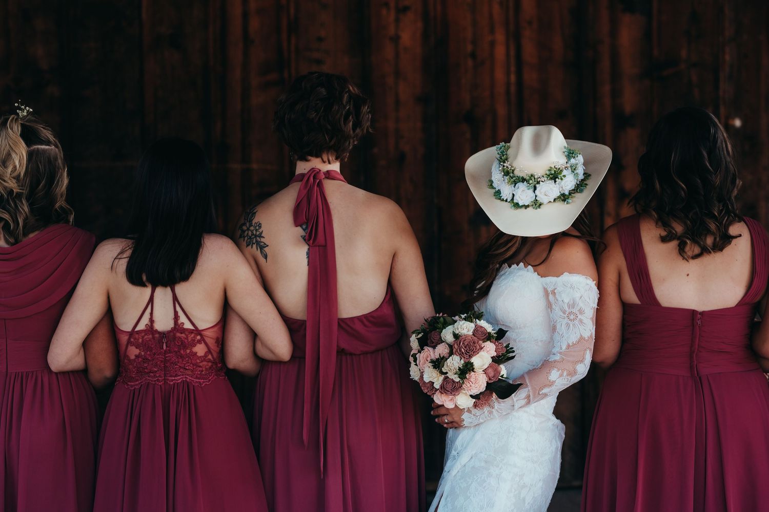Wedding party in burgundy dresses and cowboy hat posing against rustic backdrop.