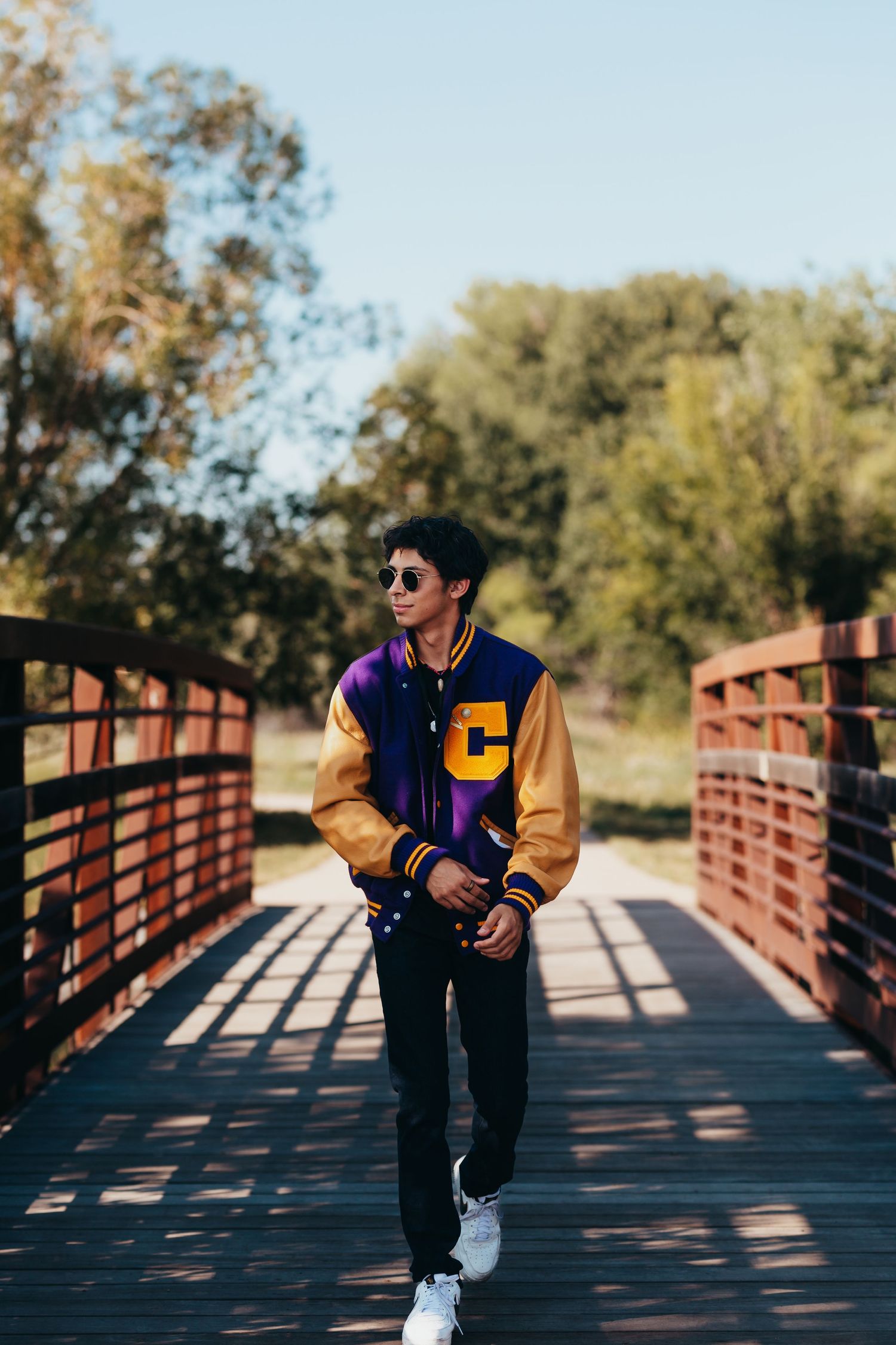 Person in varsity jacket and sunglasses posing on wooden bridge in autumn setting.