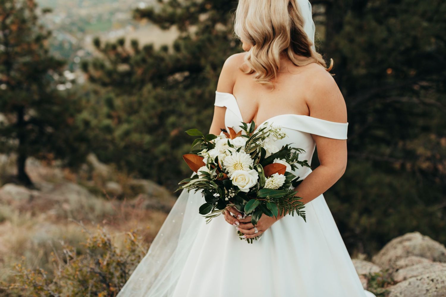 A bride in an off-shoulder wedding dress holds a lush bouquet of flowers against a mountain backdrop.