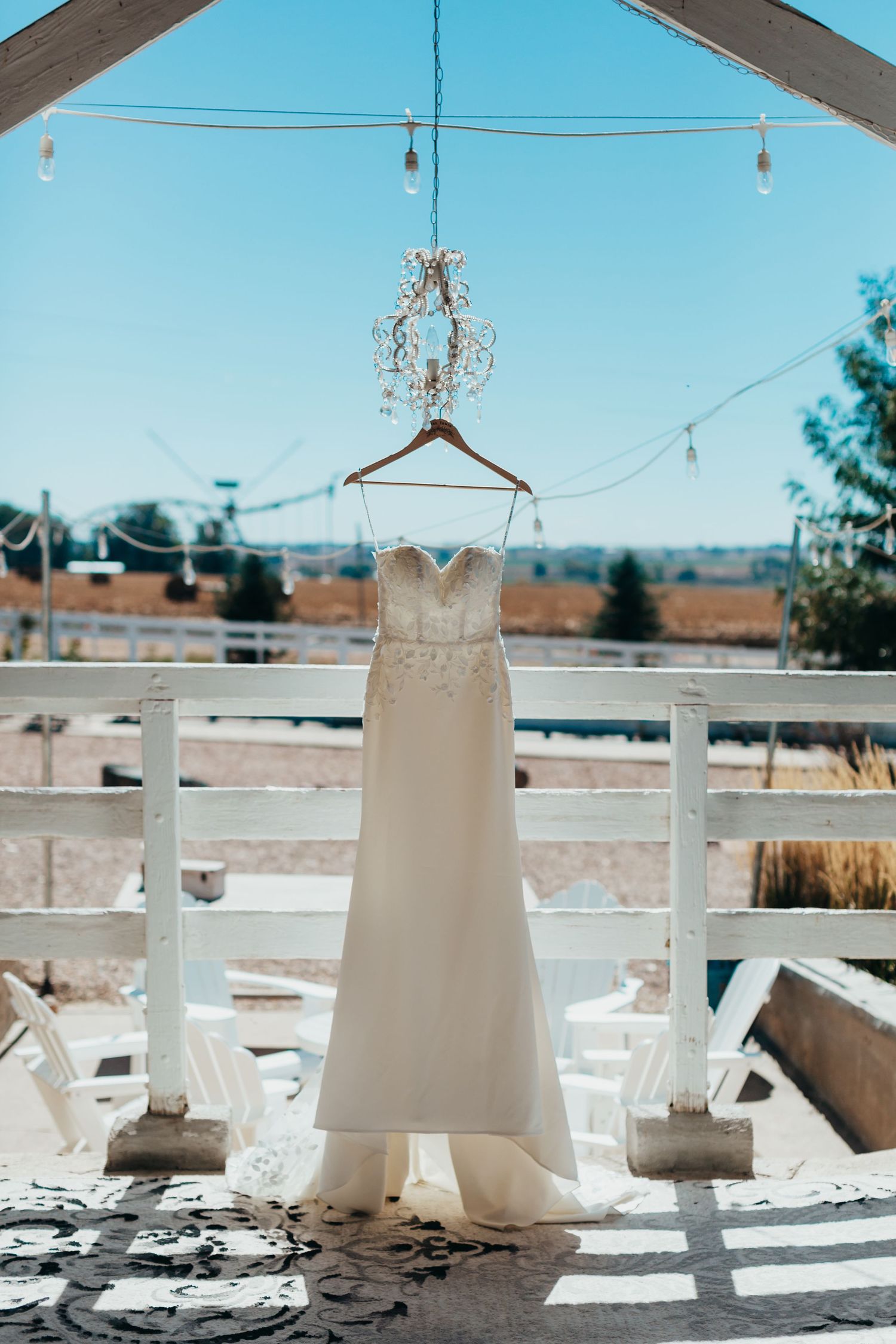 White wedding dress hanging outdoors under string lights and crystal chandelier on a sunny day.