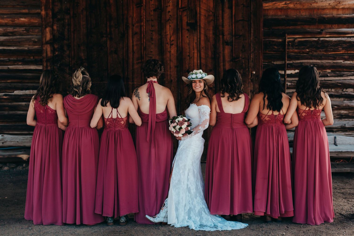 Bridal party in burgundy dresses lined up against wooden barn wall.