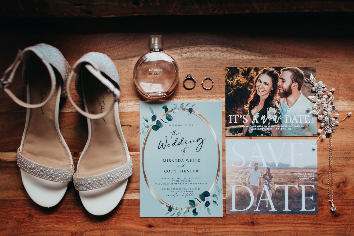 Wedding details including white shoes, invitation, perfume bottle and rings on wooden surface.