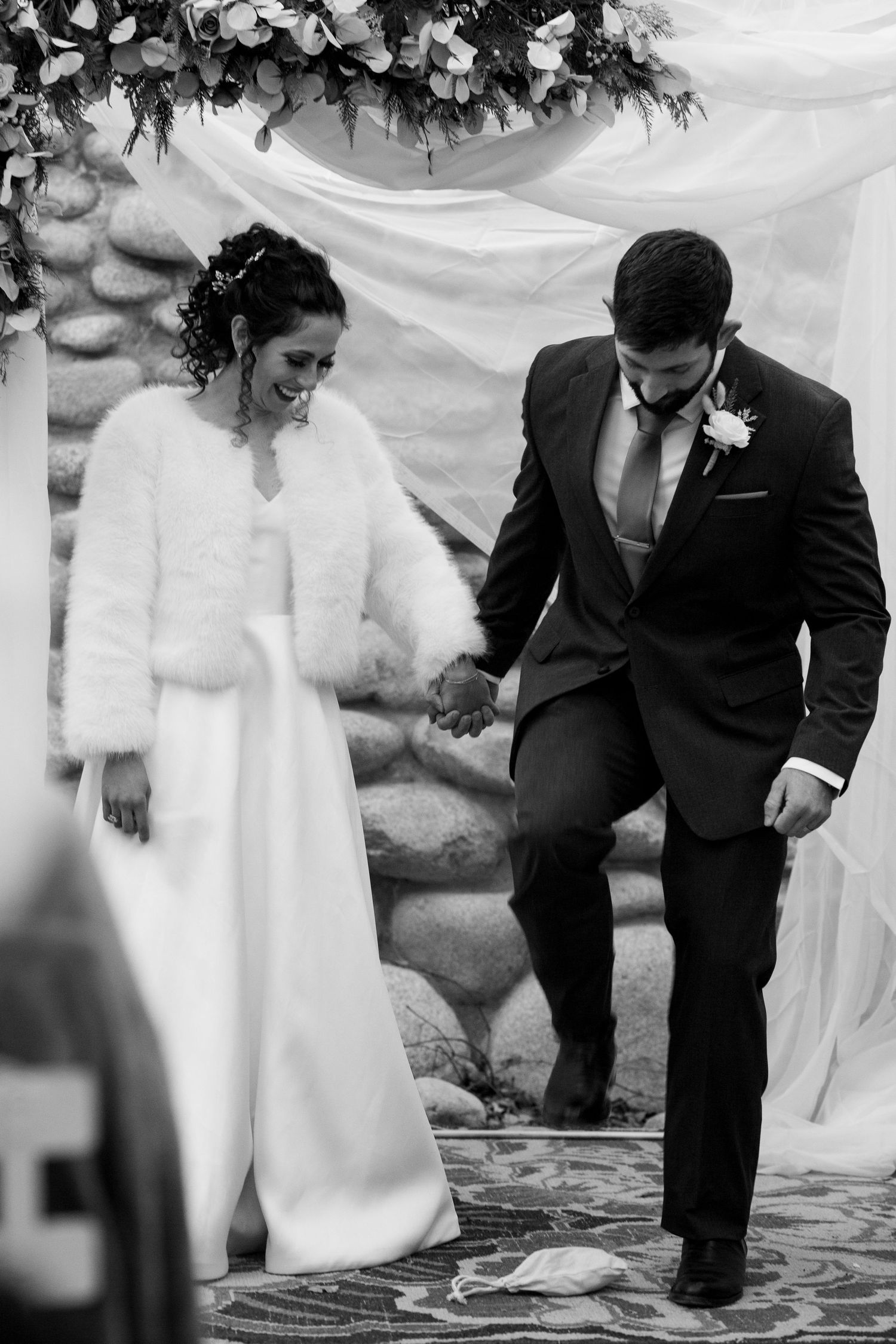 A winter wedding couple descends stone steps together while wearing classic white dress and black suit beneath floral decor.