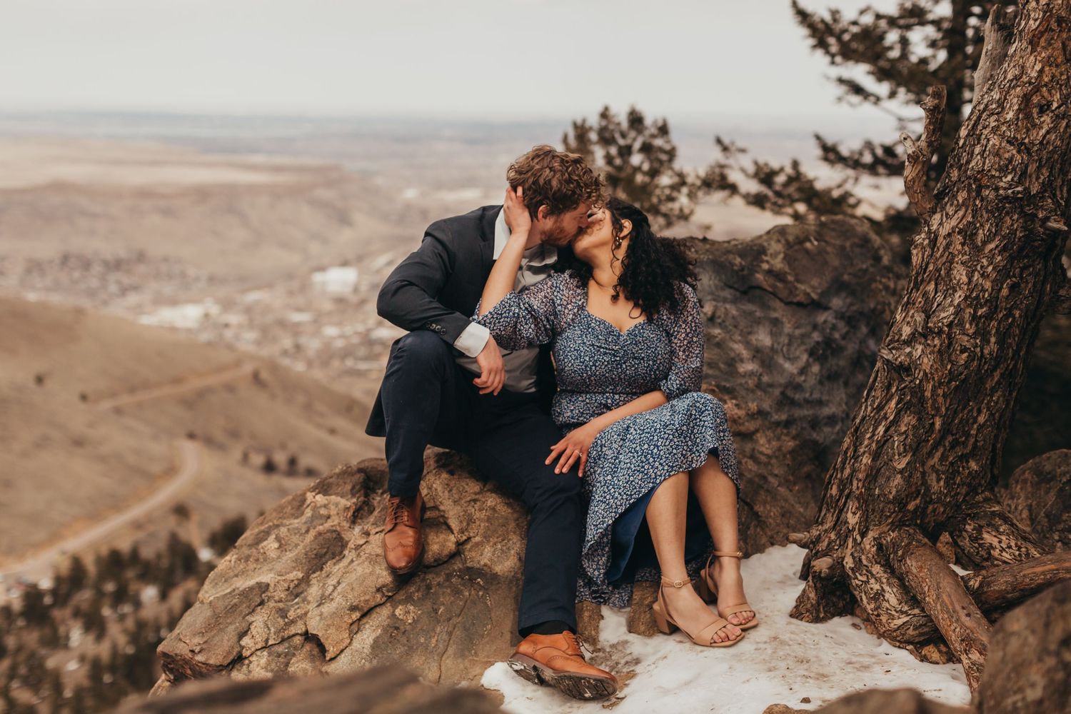 A couple sitting together on a rocky cliff overlooking a vast valley landscape.