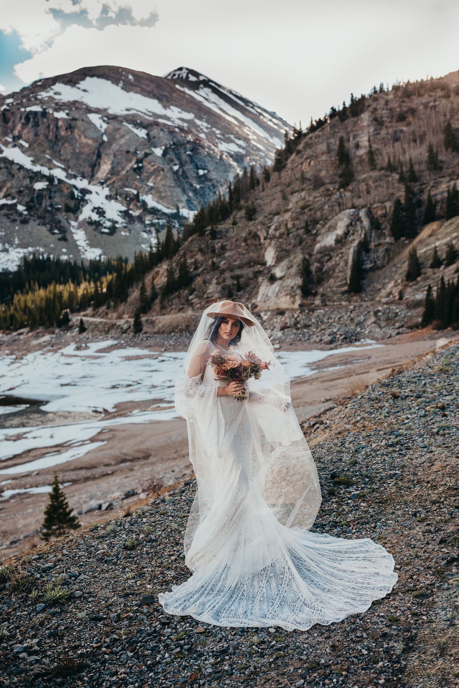 Bride in lace dress with flowing train poses against majestic mountain landscape.