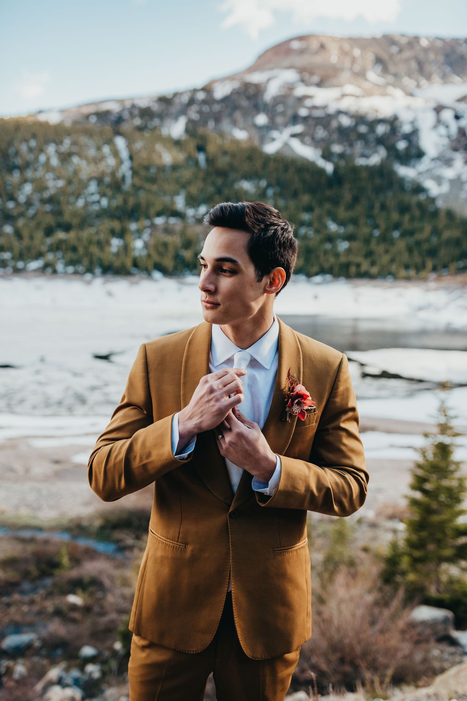 Elegant groom wearing mustard suit jacket stands before snow-covered mountain vista.