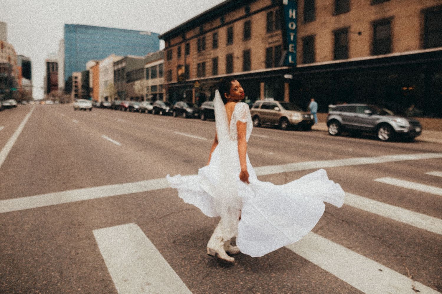 A flowing white wedding dress swirls dramatically as someone poses on a crosswalk in an urban street setting.