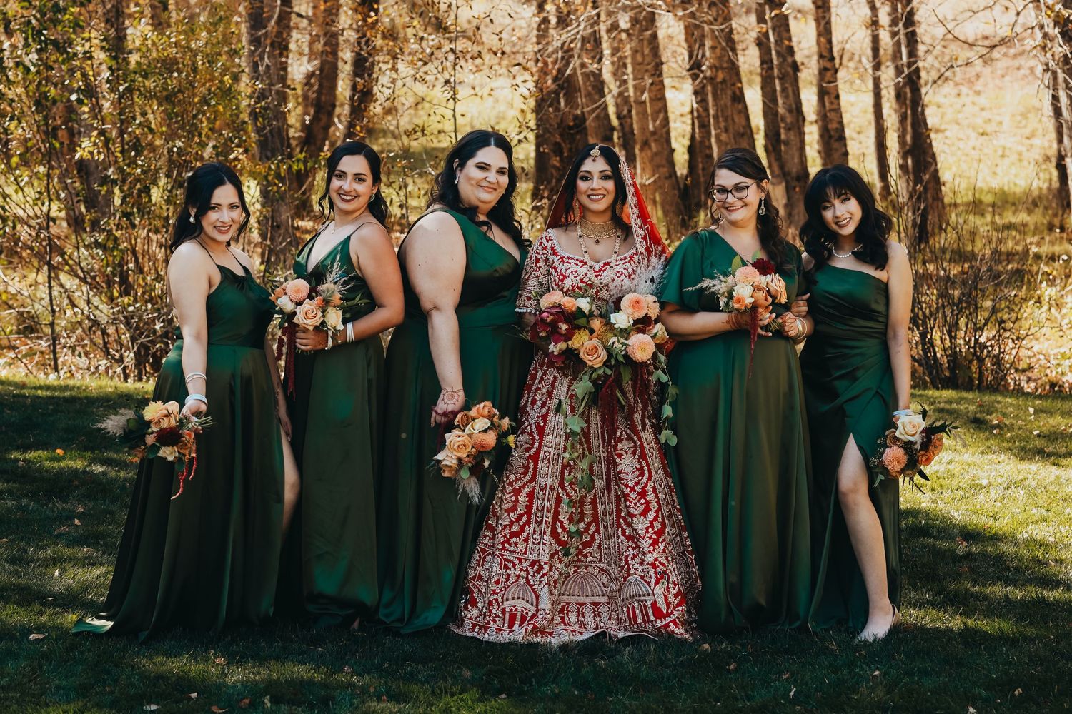 Bridal party in emerald dresses poses with bride in red lehenga during fall wedding.
