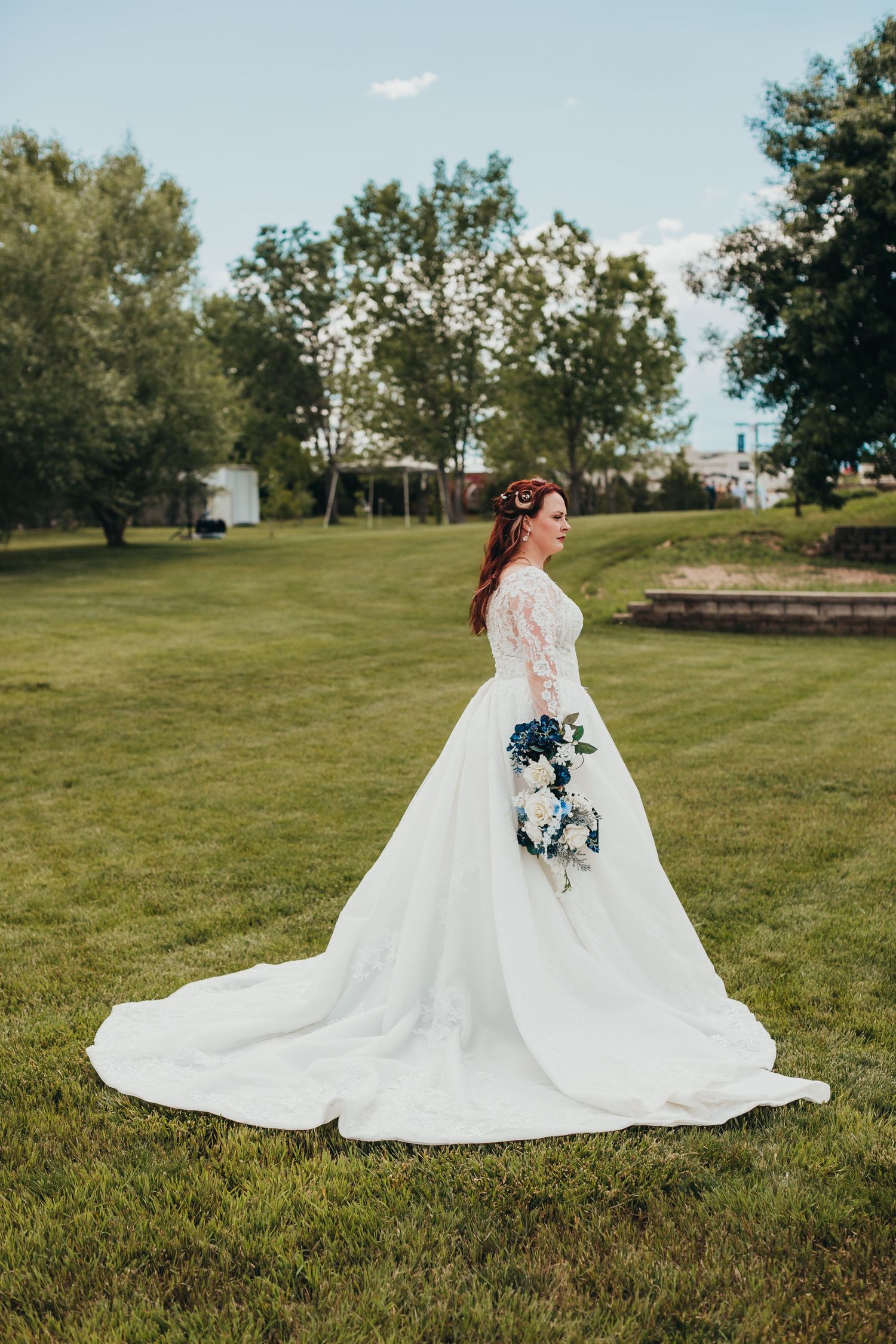 Side view of bride in elegant white gown holding blue bouquet in sunny garden setting.