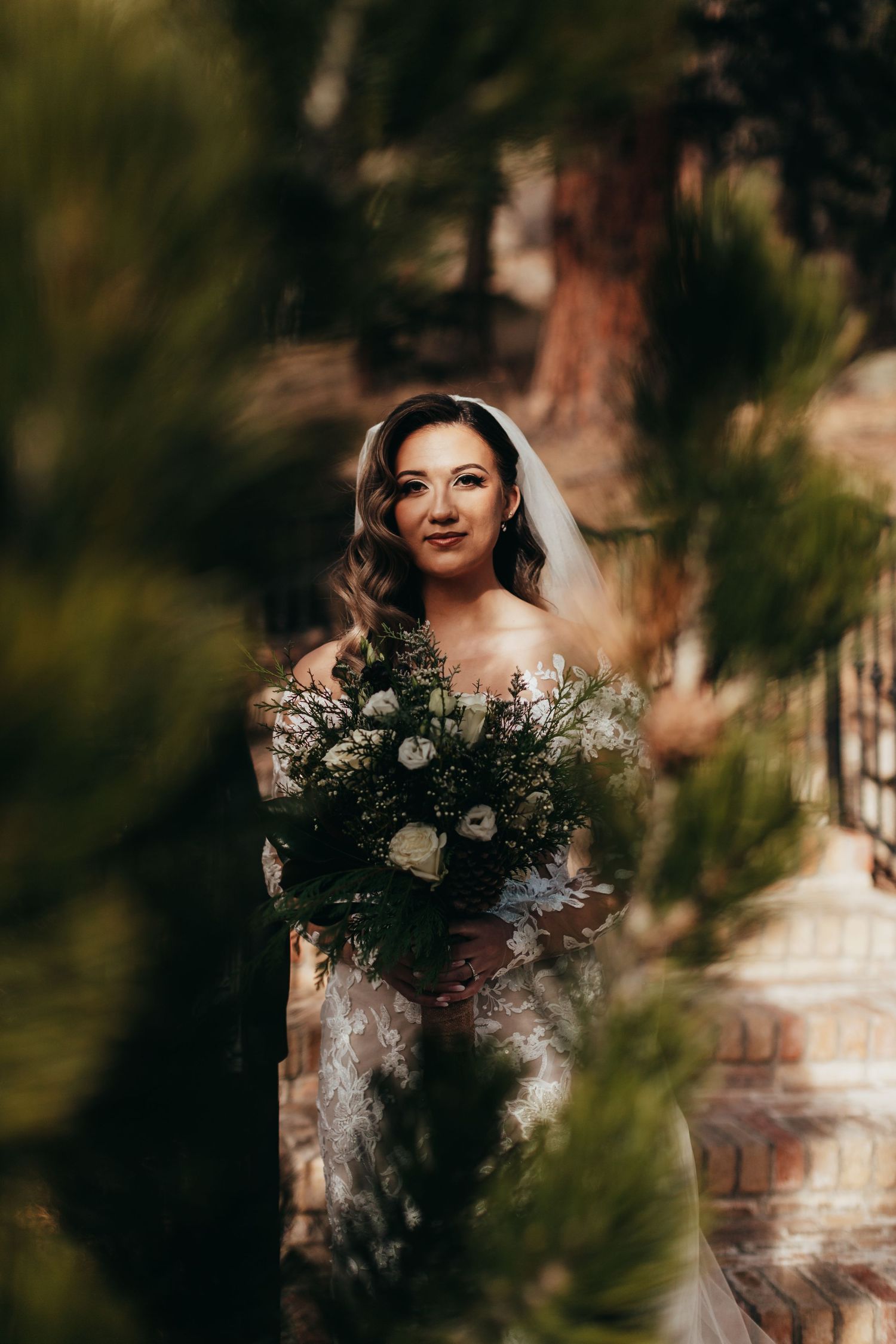 A bride in a white lace gown holds a large neutral-toned bridal bouquet in a dreamy outdoor setting.