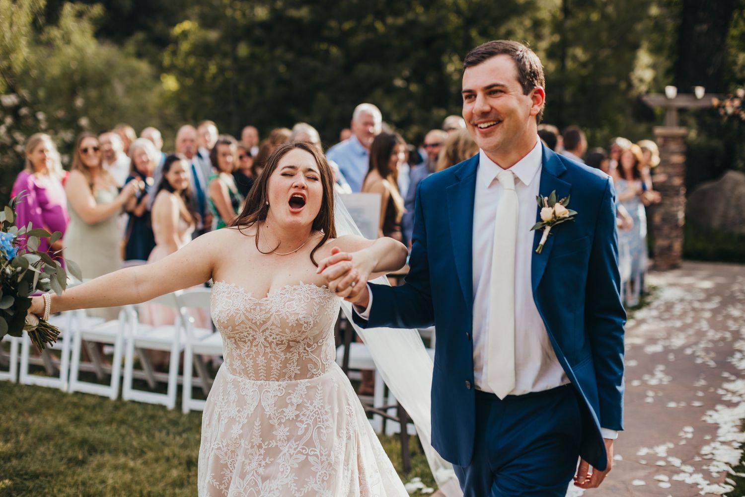 Newlyweds celebrate with joyful expressions while walking down outdoor aisle.
