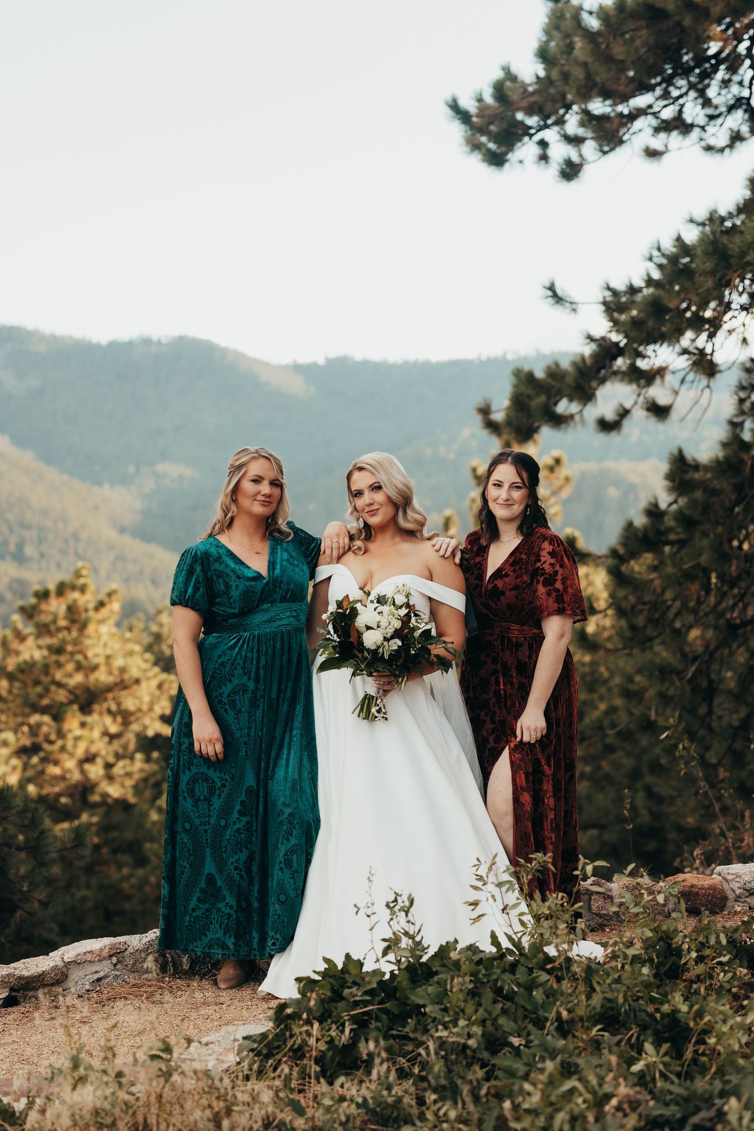 Three women in elegant dresses pose on a mountain overlook during a wedding celebration.