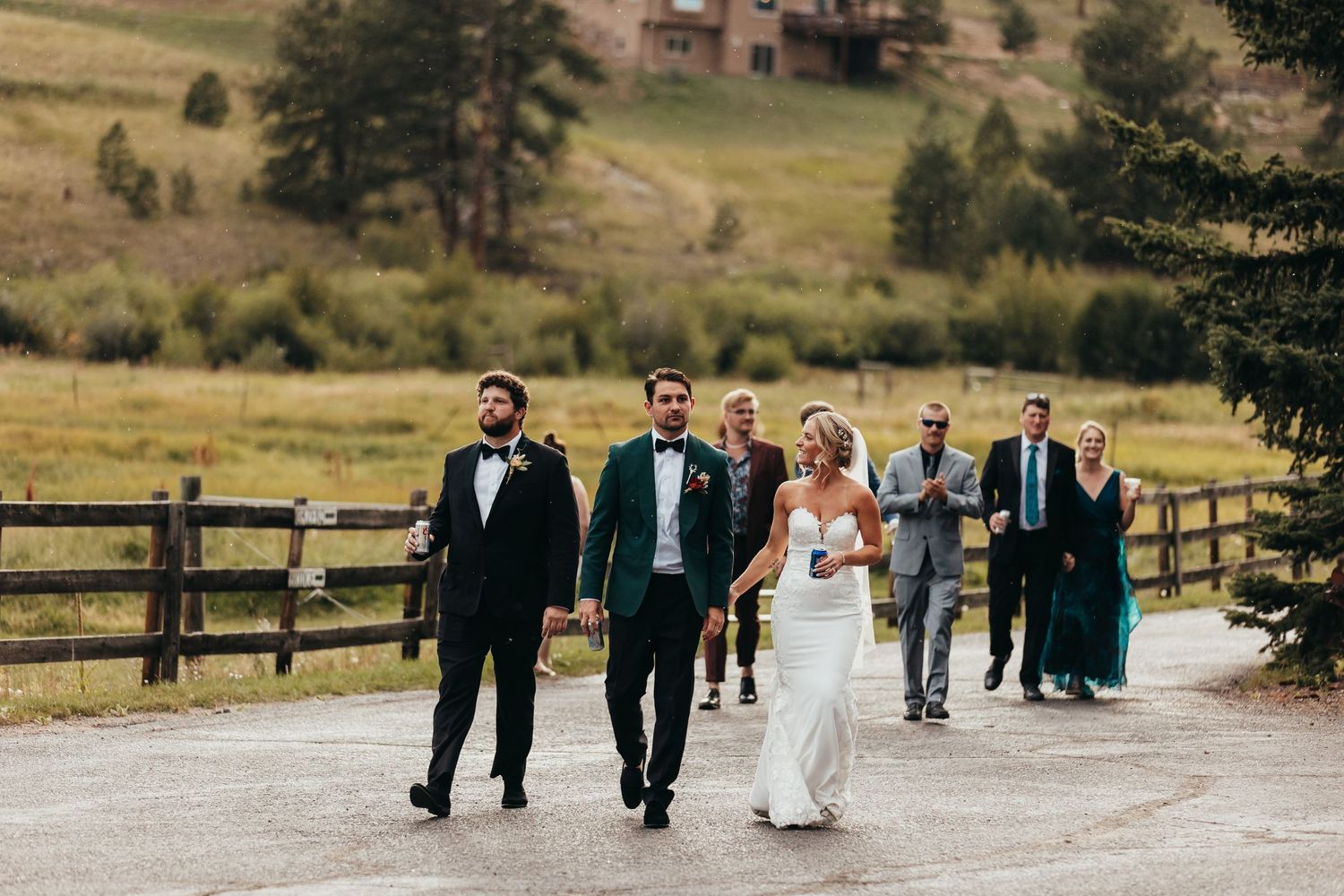 Wedding party walks along a path with mountain scenery and wooden fencing in the background.