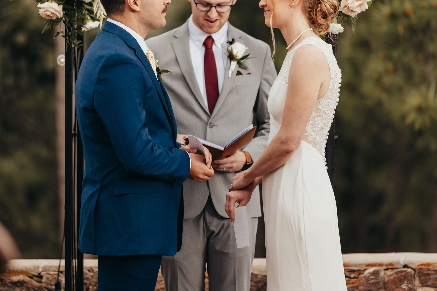 Wedding ceremony outdoors with officiant in gray and couple exchanging vows under an arch.