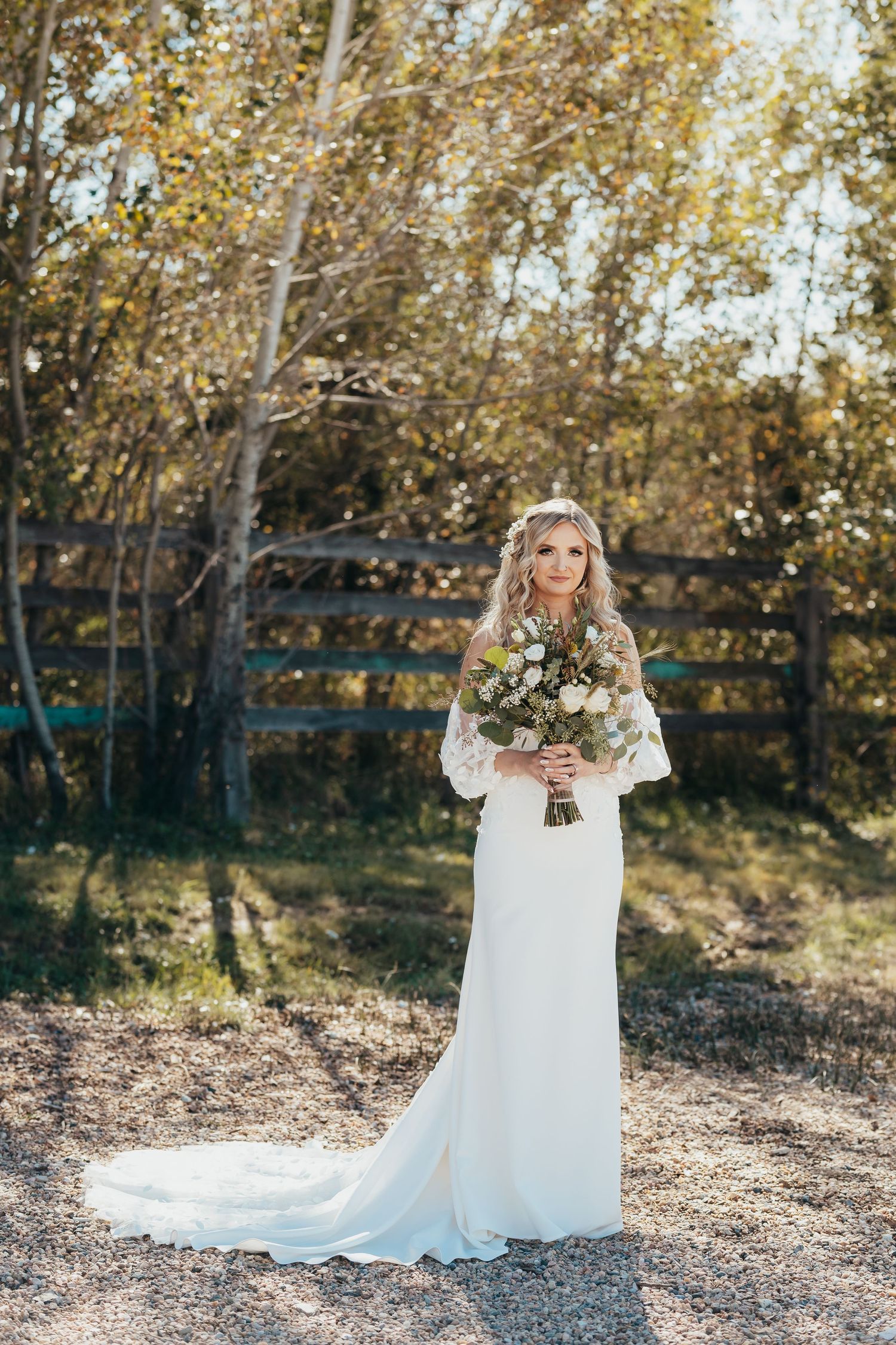 Bride in fitted white dress with lace sleeves holds rustic bouquet against autumn forest backdrop.